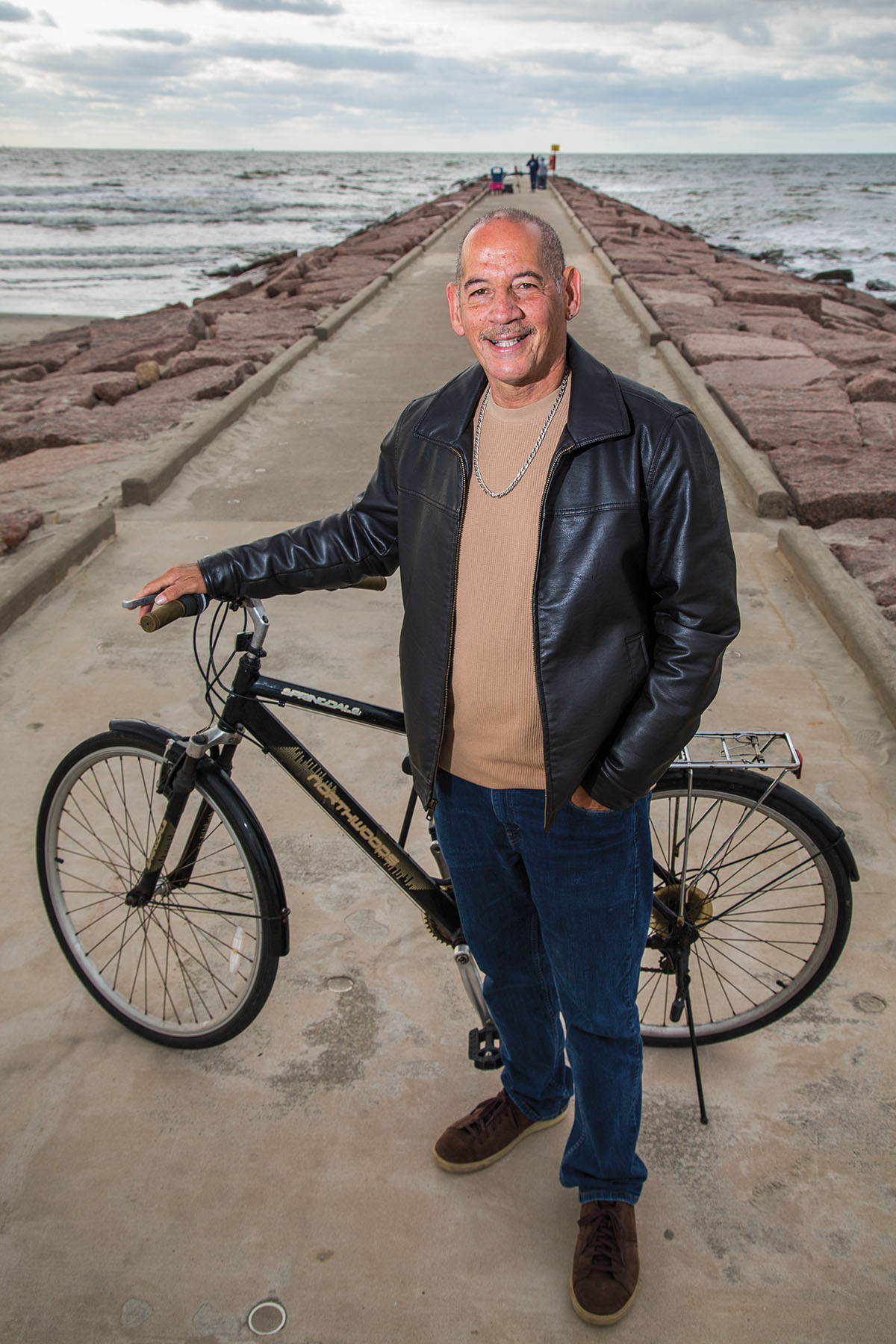 A man in a leather jacket and tan t-shirt stands on a pier holding a bicycle