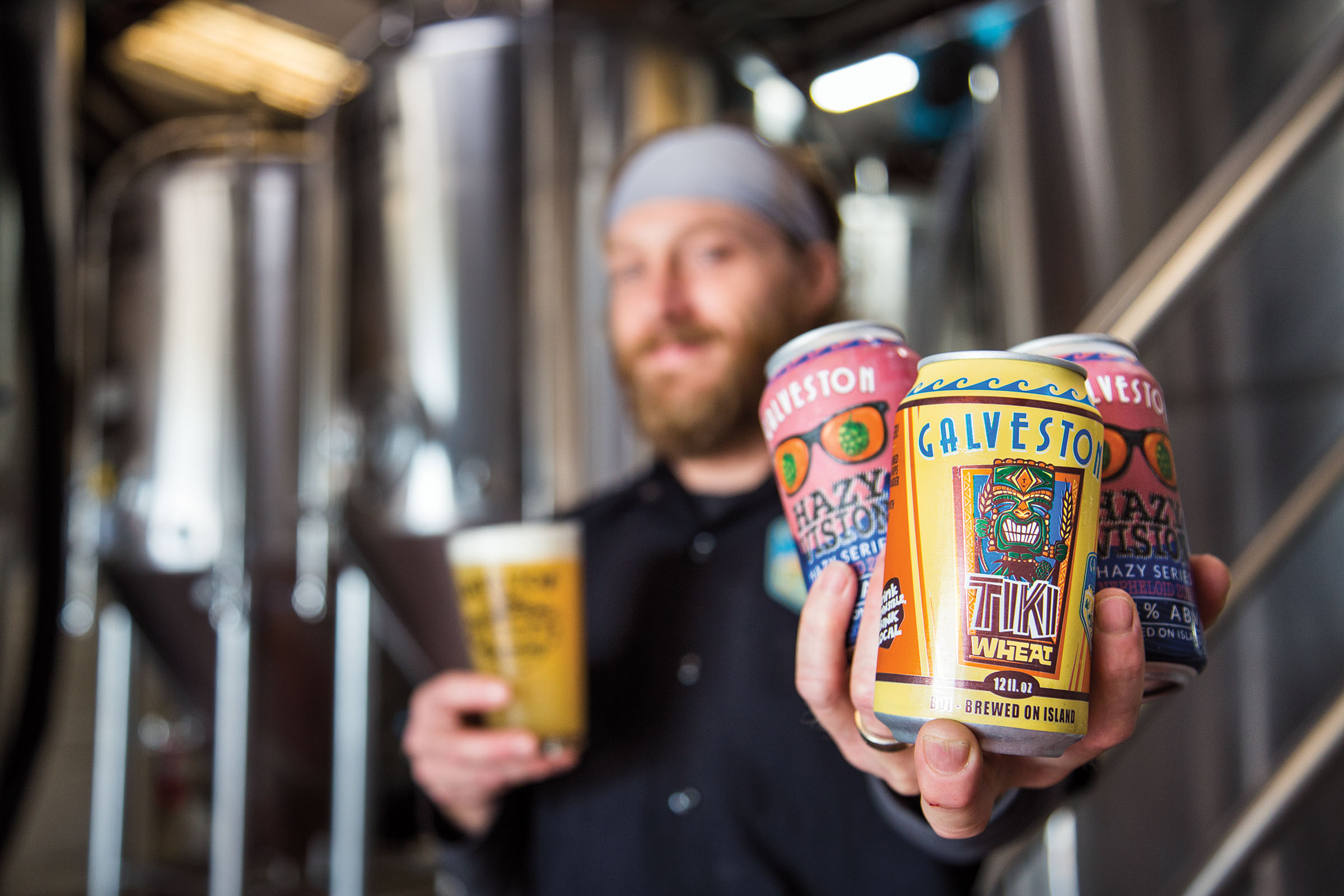 A man holds three brightly-colored cans of beer in his hand in front of brewing equipment