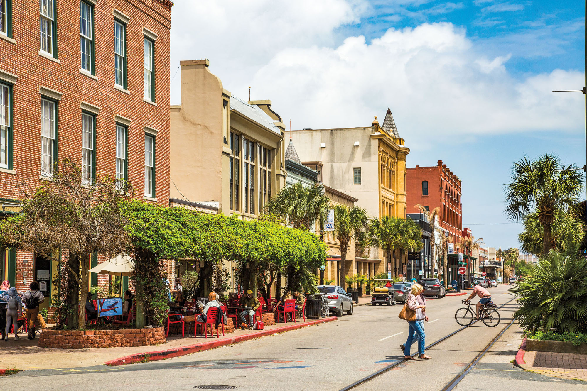 Old brick buildings and green trees line a street under blue sky