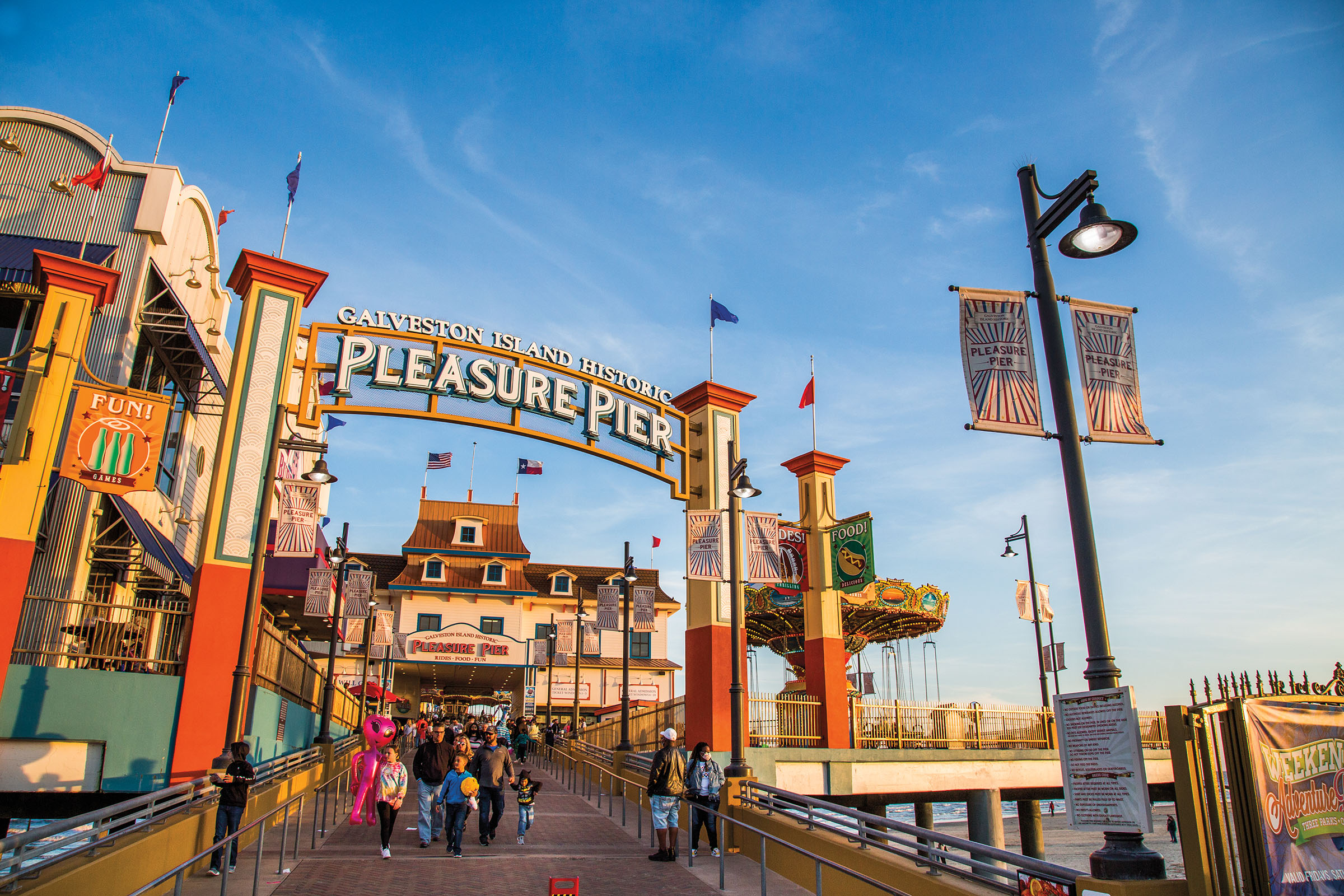 Beautiful blue sky and red and blue flags adorn a walkway with a sign "Galveston Island Historic Pleasure Pier" with people walking underneath