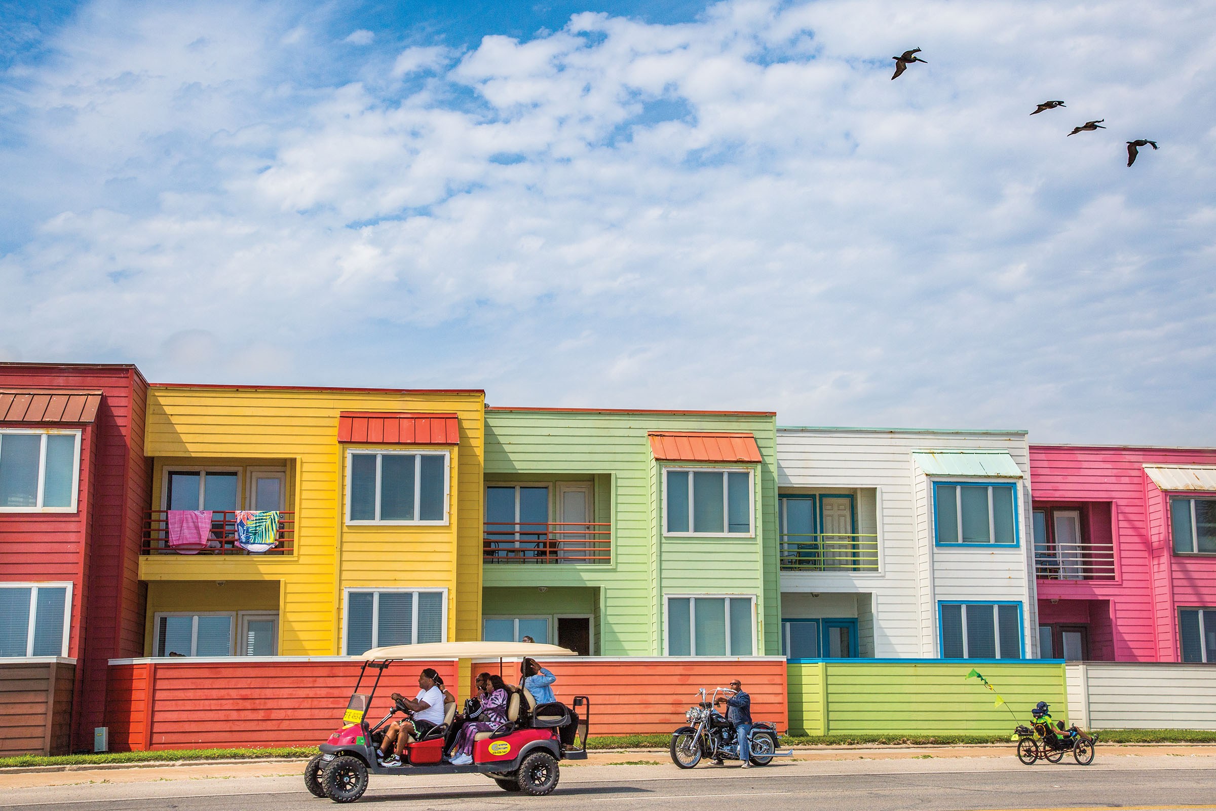 A small group of people in golf carts and motorcycles drive in front of brightly colored buildings under a blue sky