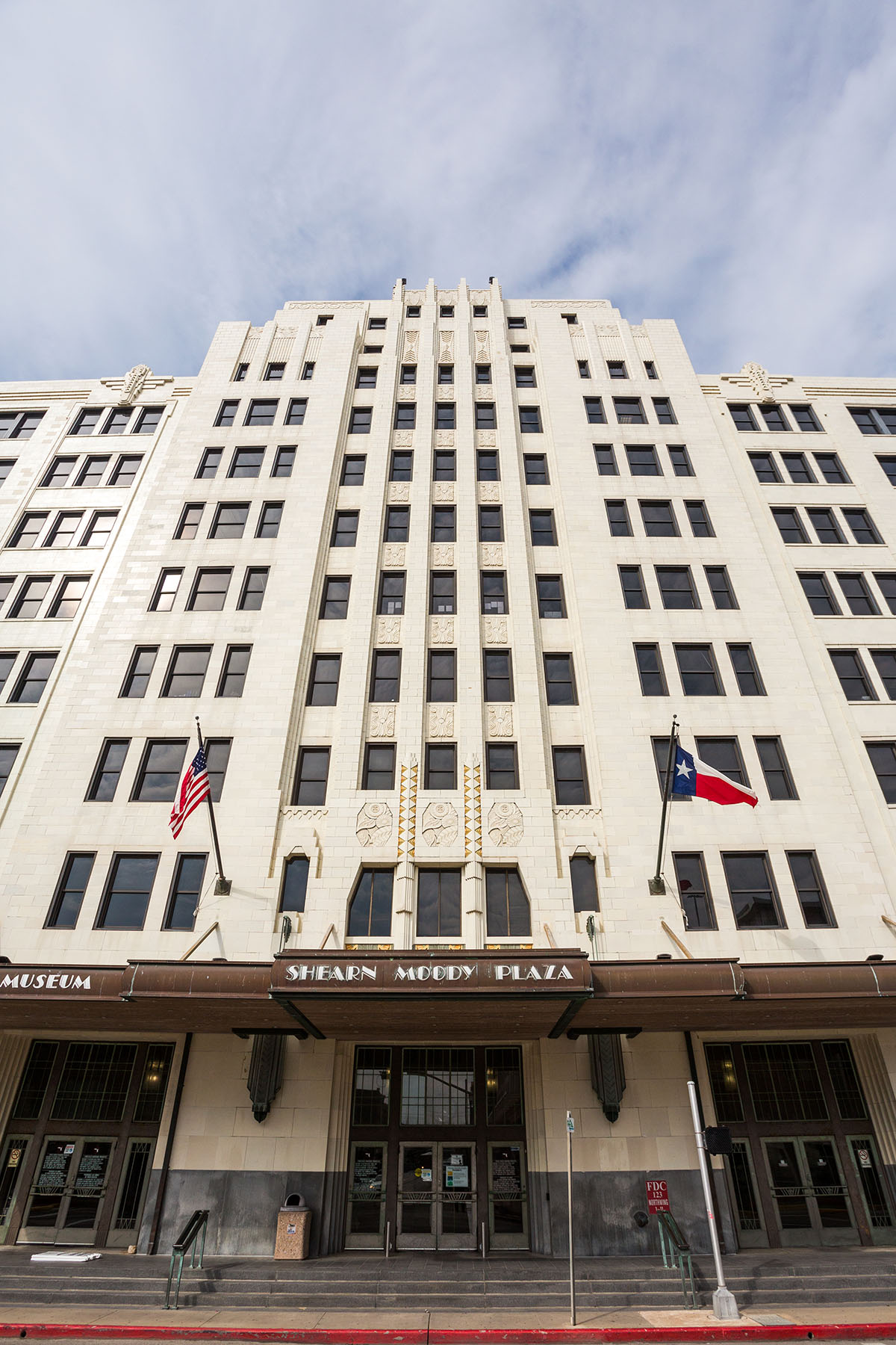 The outside of a tall, white building with a U.S. and Texas flag and a sign reading "Shearn Moody Plaza"