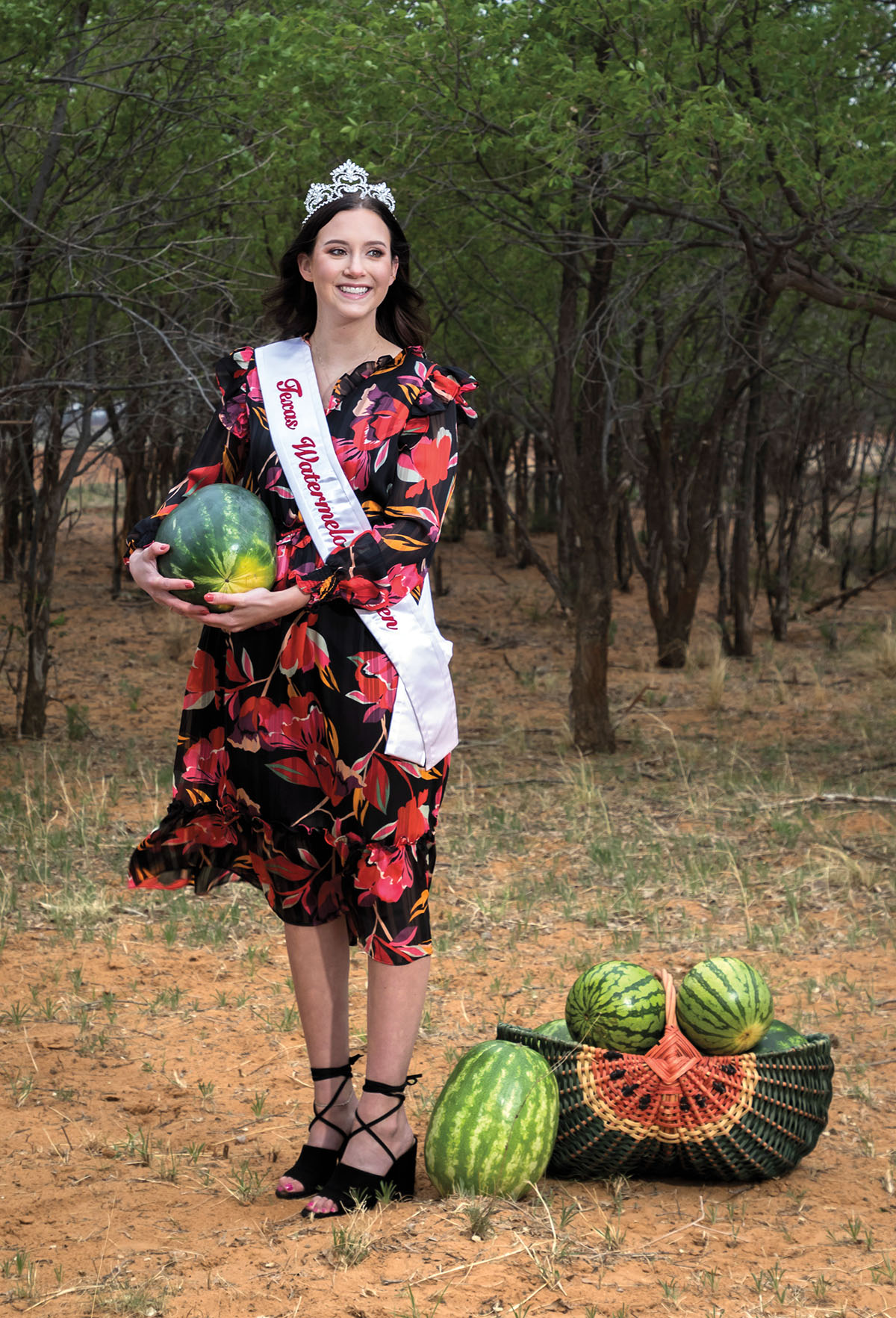 A woman in a flowered dress and sash holds a watermelon standing next to a basket of watermelons