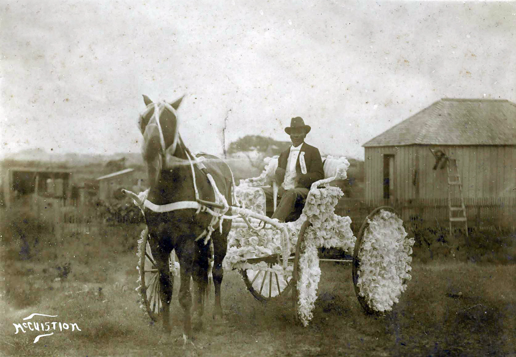 A man in a suit and hat sits in a decorated horse carriage behind a horse in a vintage photograph
