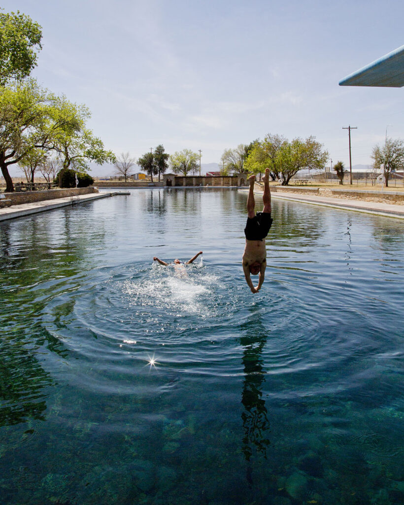 A person jumps off of a diving board to meet another swimmer already in the clear blue water of Balmorhea
