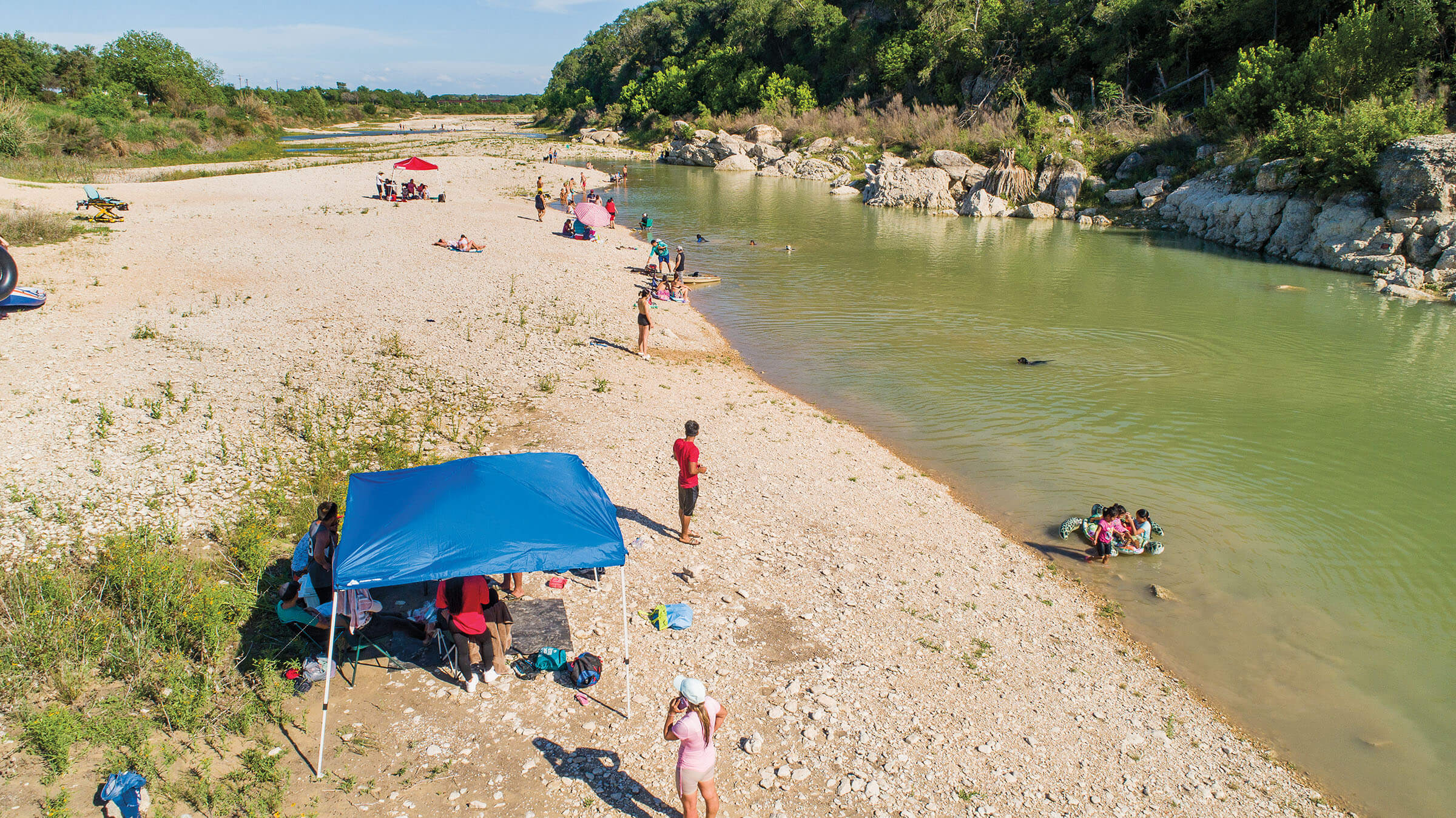 Groups of people sit under tents or in the sun along the banks of a blue-green river