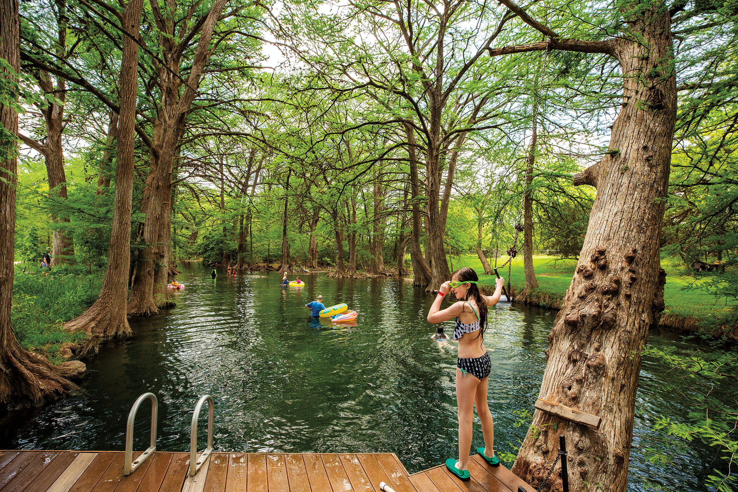 A young woman in swim goggles stands on a wooden dock on the edge of the blue Blanco river water