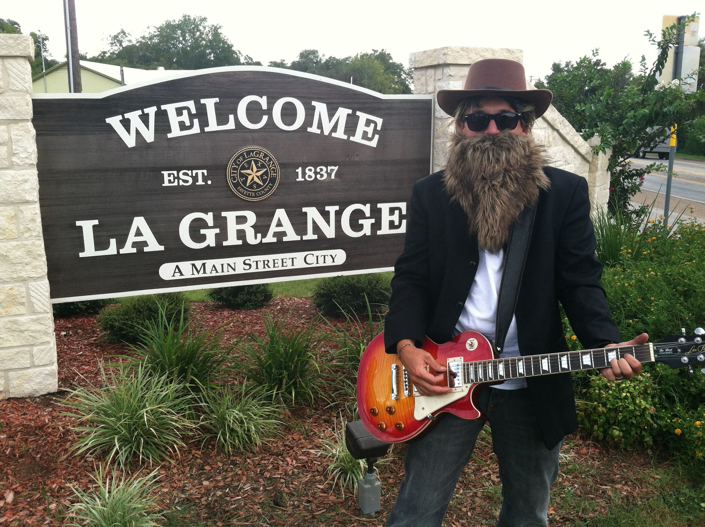 A man wearing a hat and false wig stands with a guitar in front of a sign reading "Welcome to La Grange"