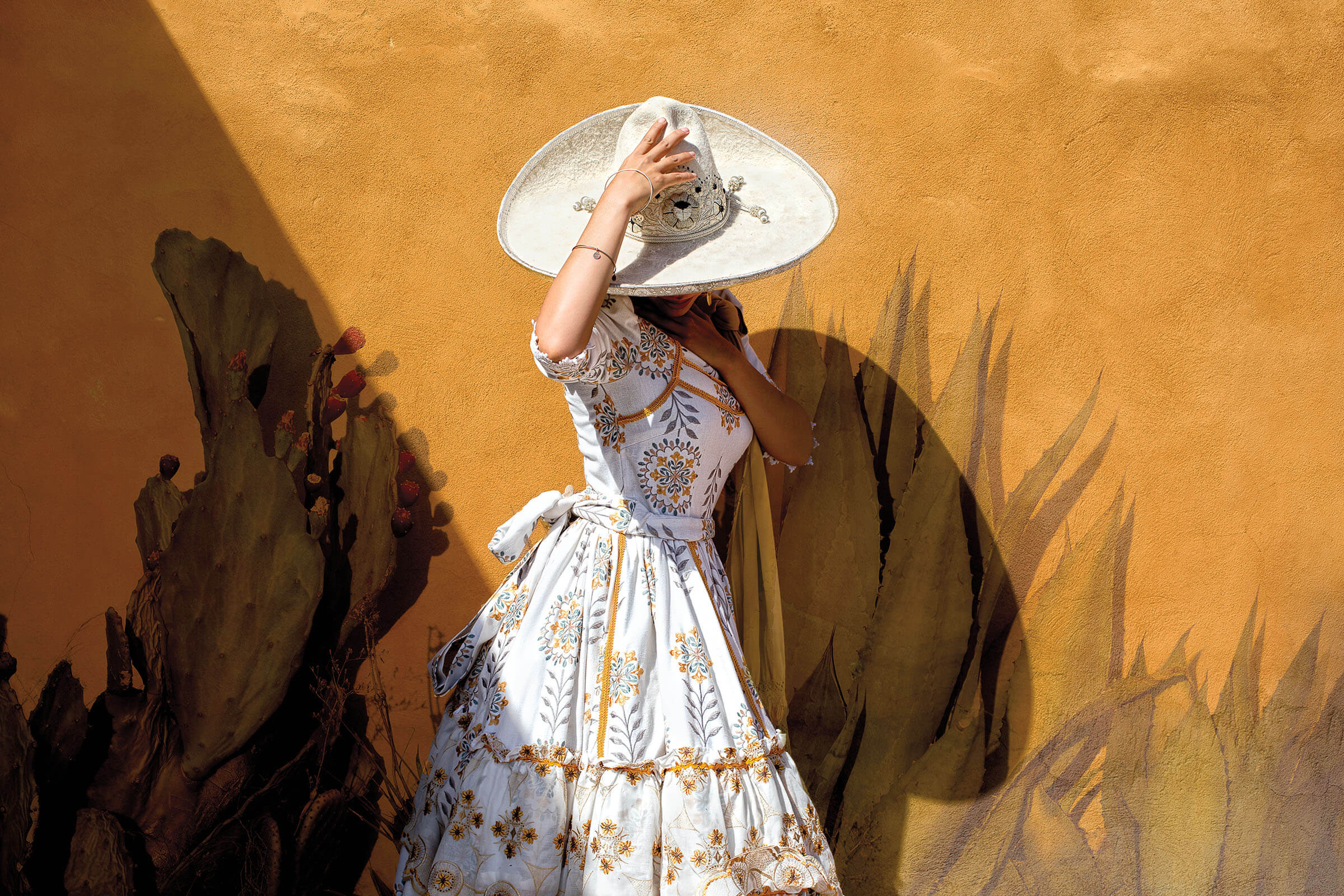 A woman in a white dress holds a large white sombrero over her head in front of a golden yellow painted wall