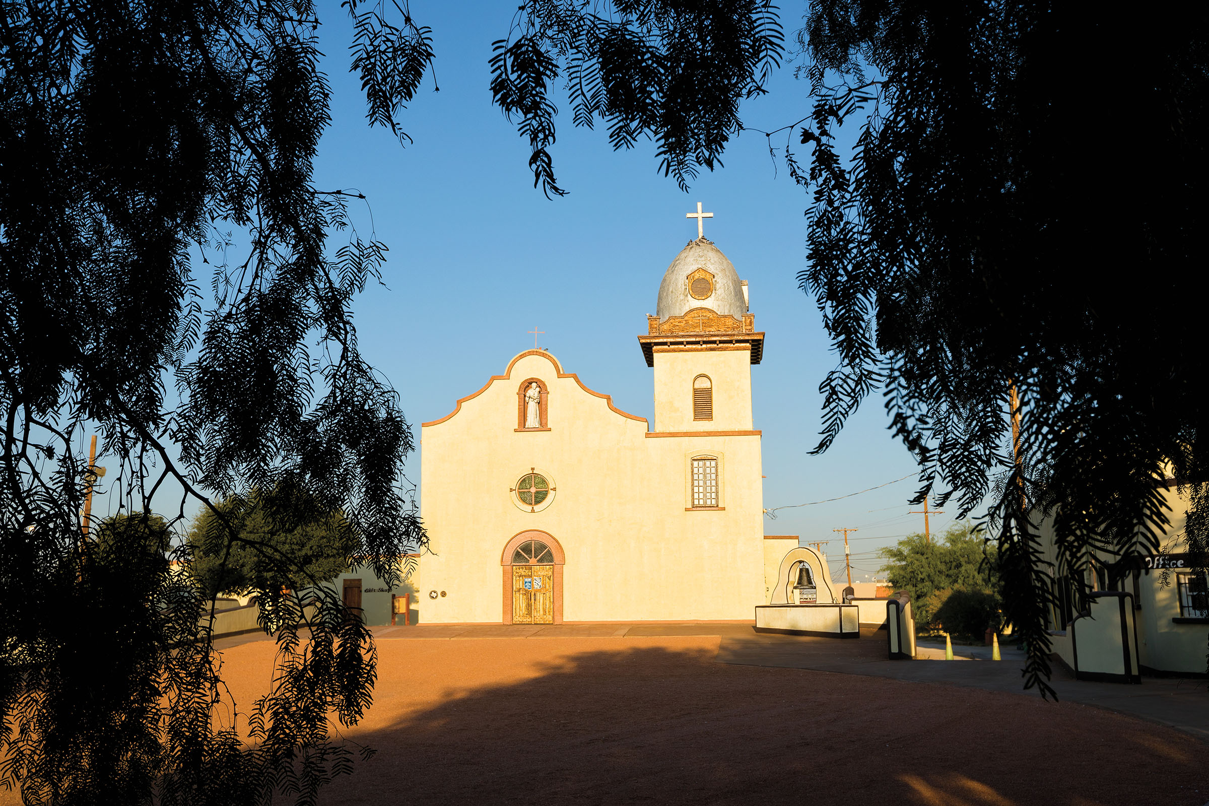 The sun shines dow on an adobe-walled mission building with golden trim and a white cross in front of a dark tree