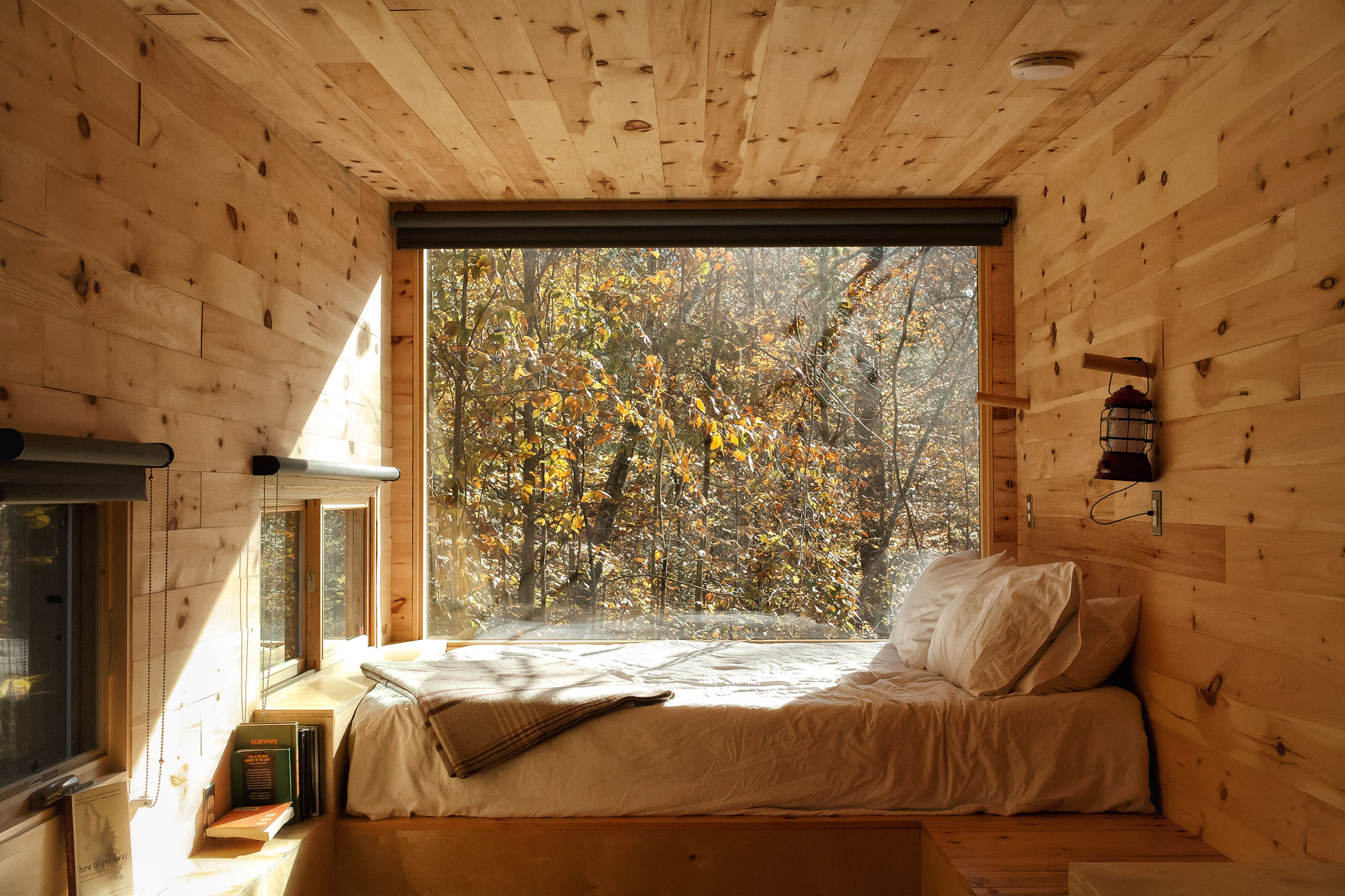 The view through a window to a lush green outdoor scene above a white bed with pine walls
