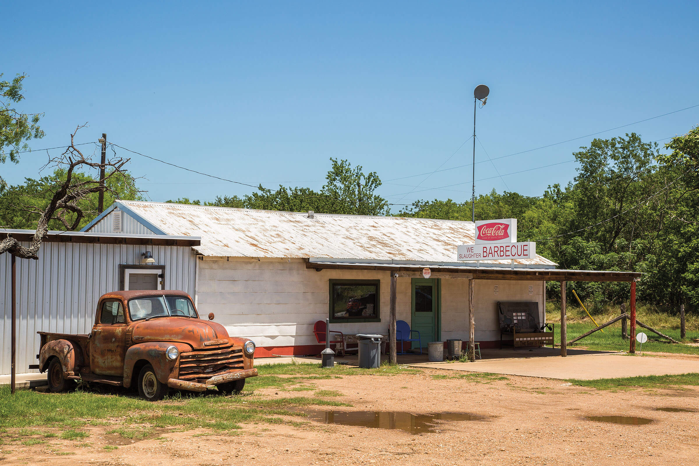 The outside of a white building with a rusting roof and a sign reading "We Slaughter Barbecue"