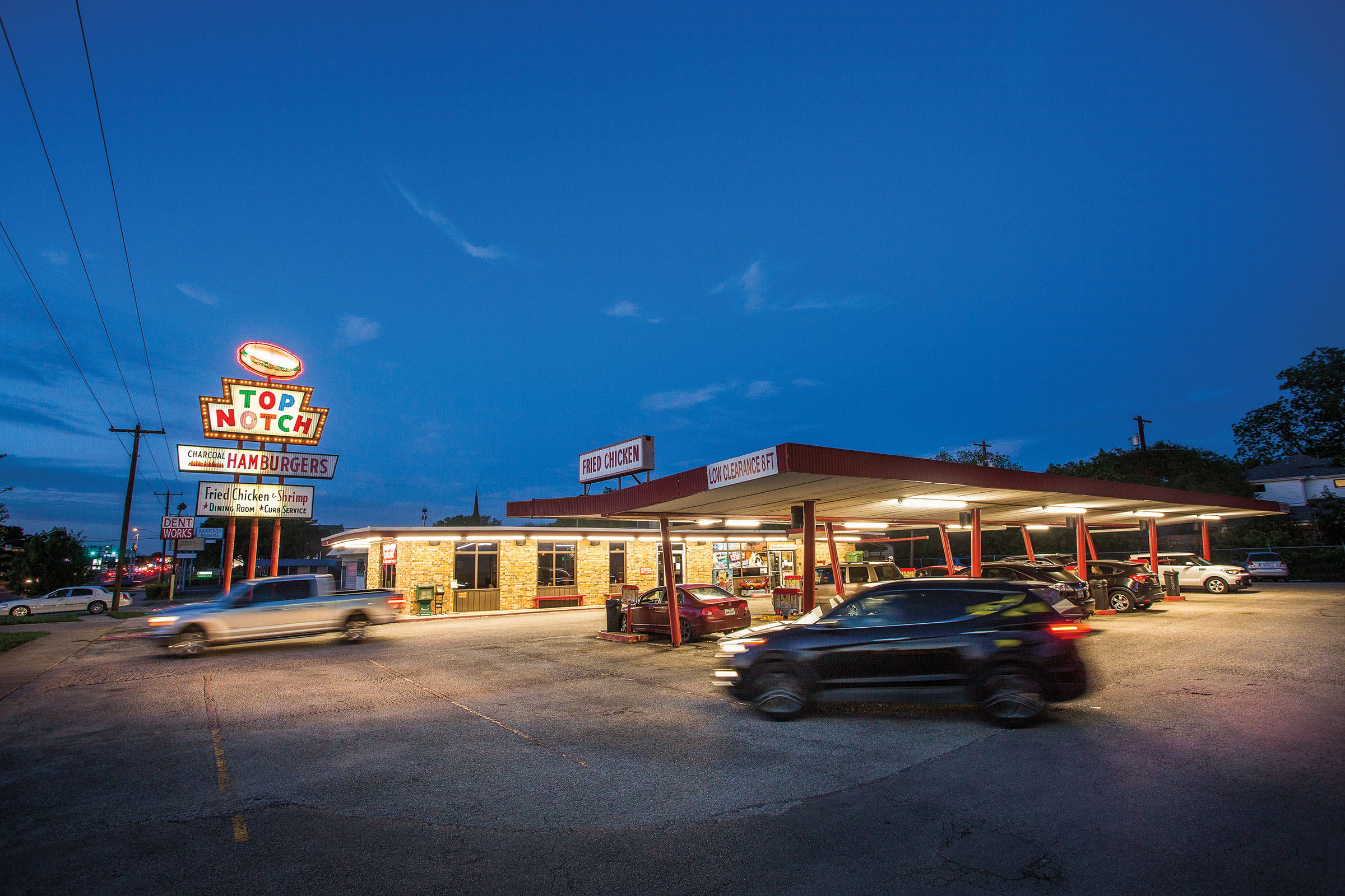 Cars drive in and out of a parking lot under a well-lit sign reading "Top Notch Hamburgers" at dusk