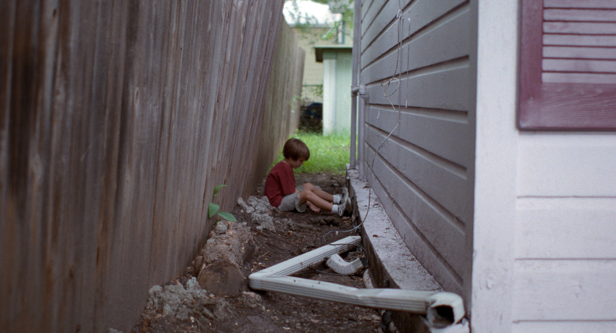 boyhood still little child sits in between house and fence in red shirt and tennis shoes