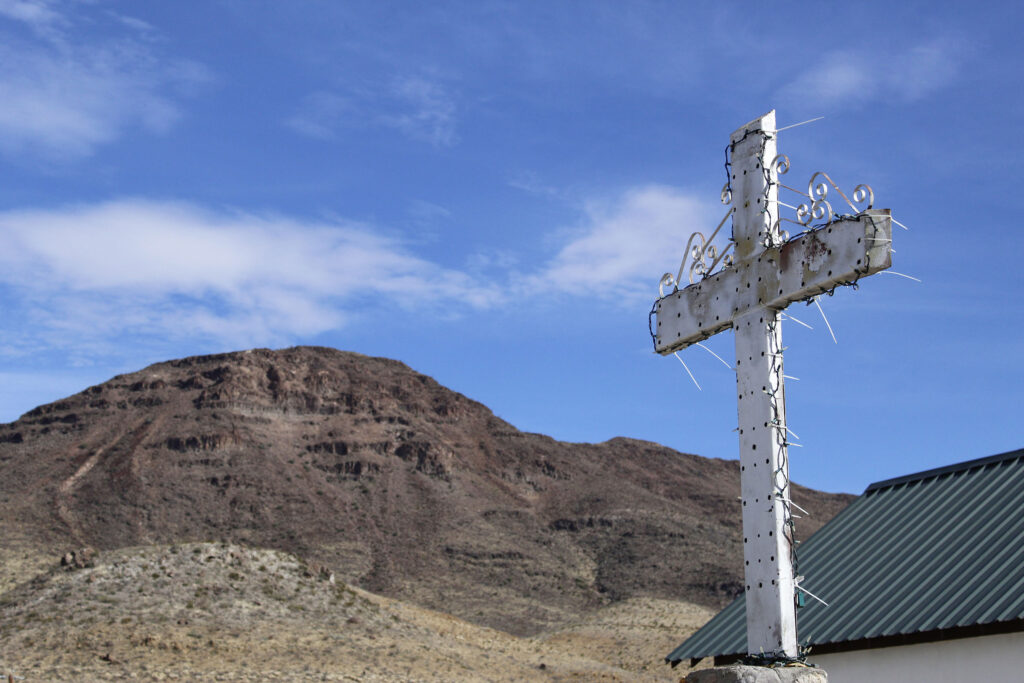 A white cross sits in the foreground of a landscape with mountains. The Sacred Heart Catholic Mission Church in Shafter, captured in a photo titled "Church." Photo by Morgan Page, courtesy of Bones of Texas. 