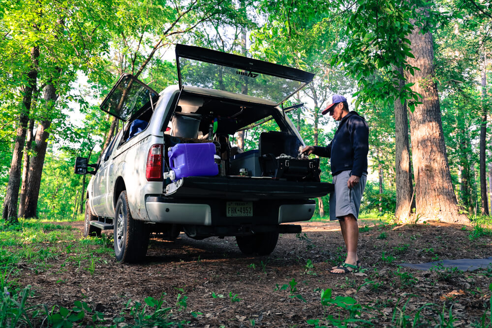 A man stands cooking on the tailgate of his truck surrounded by tall green trees
