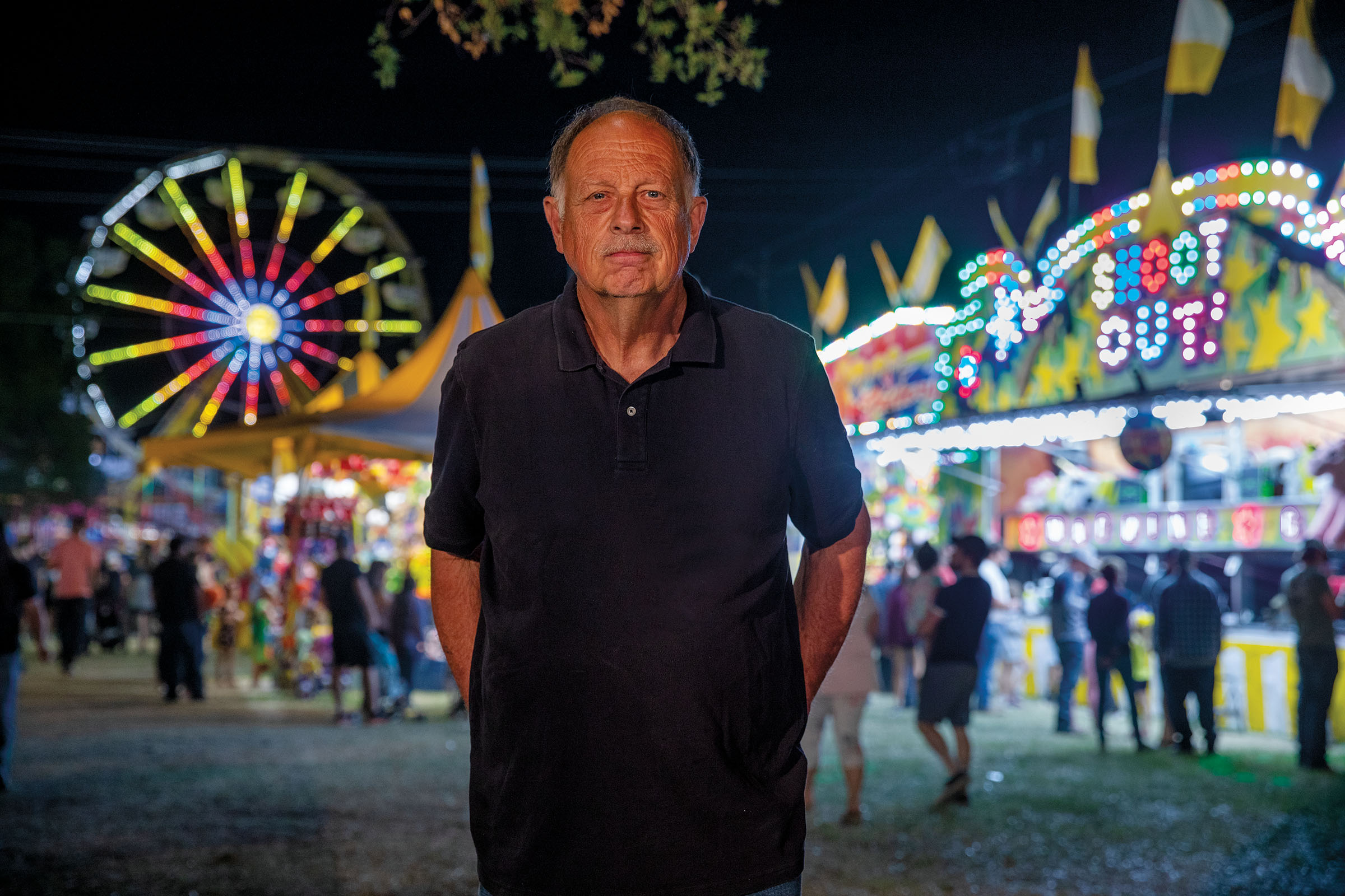 A man in a blue polo shirt stands in front of brightly colored, lit-up carnival rides