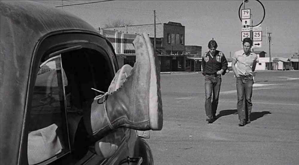 A black and white screen capture shows a foot sticking out of a car window with two young men walking nearby