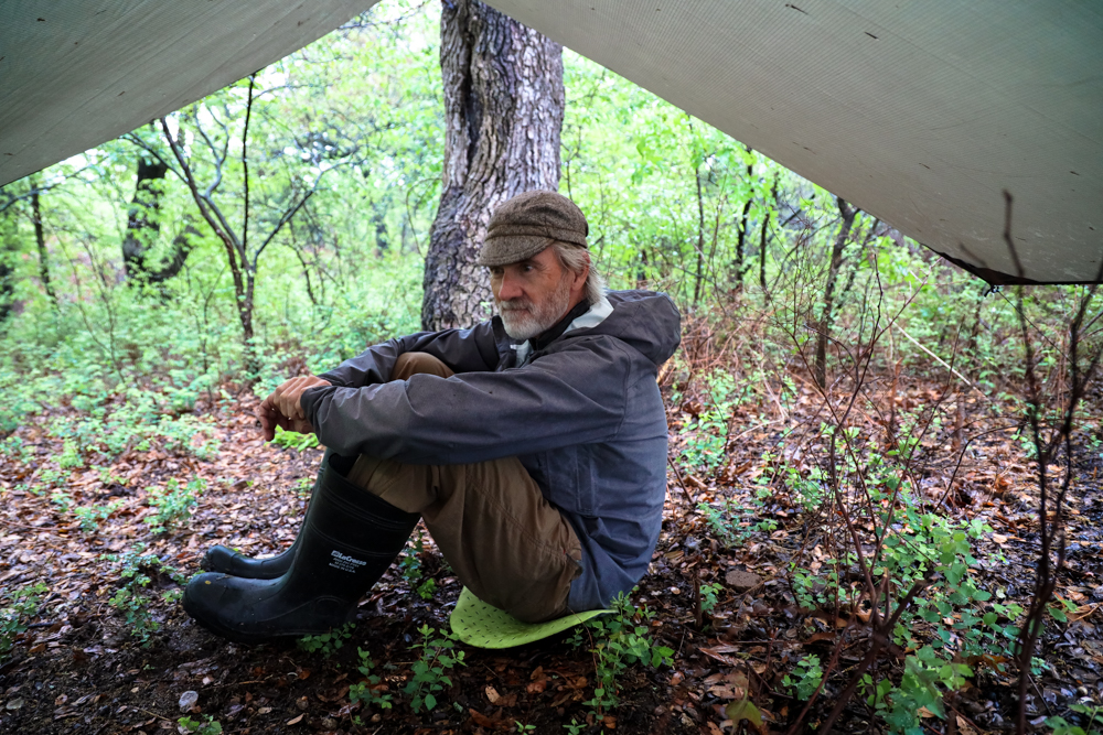 A man in a baseball cap sits under a tarp grabbing his knees