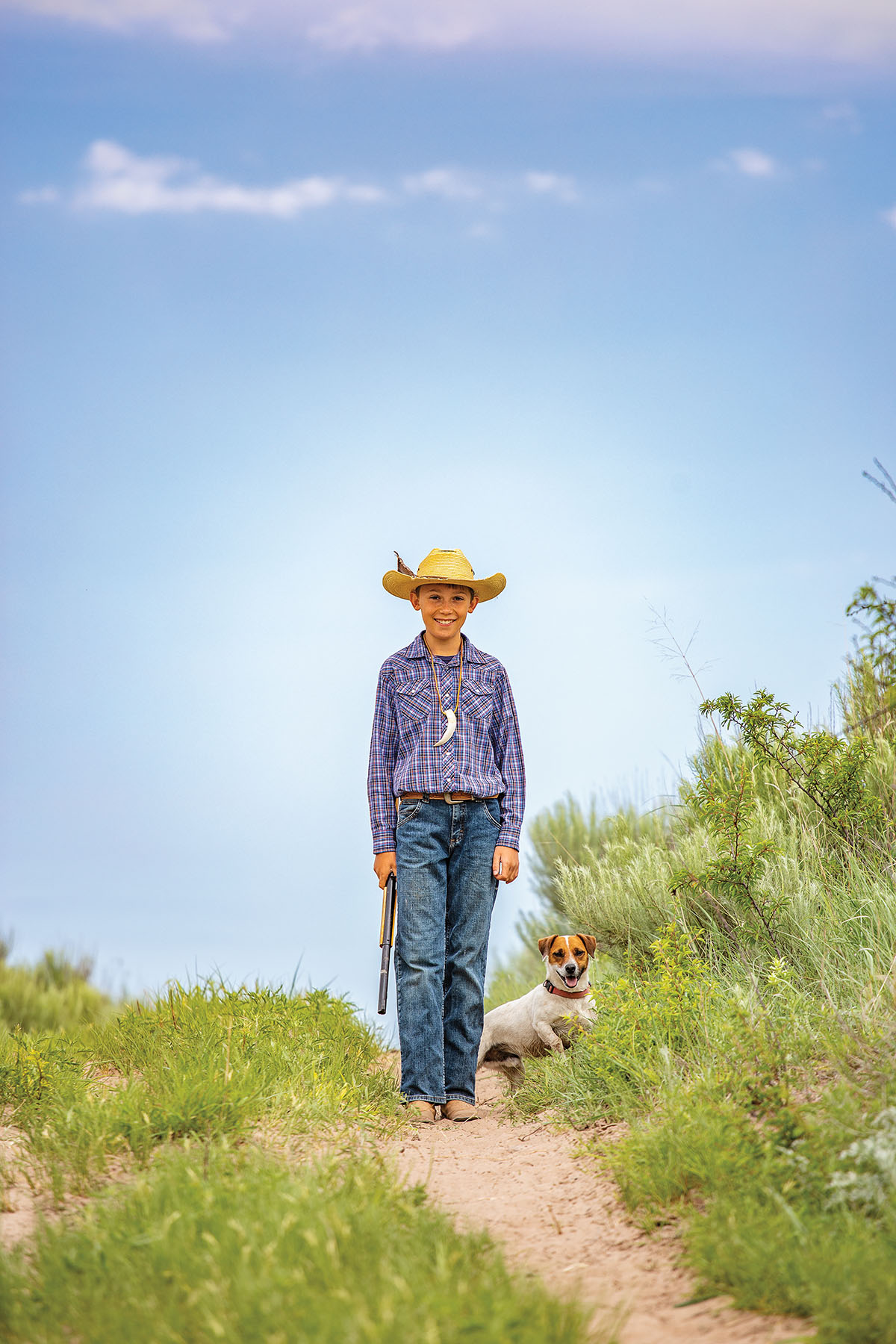 A young man in a collard shirt and cowboy hat walks down a dirt trail with a dog by his side