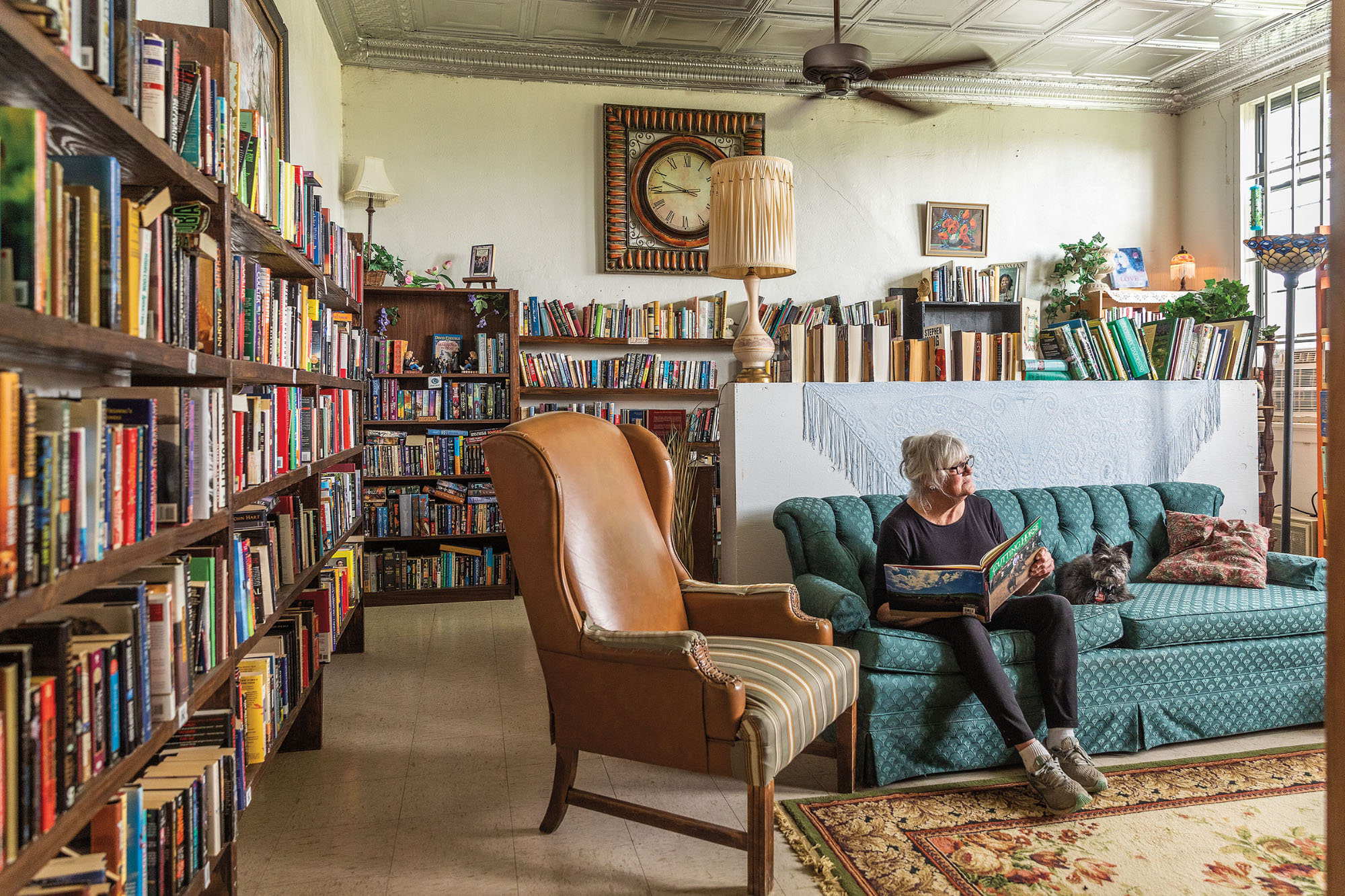 A woman sits on an old green couch next to a leather chair surrounded by books