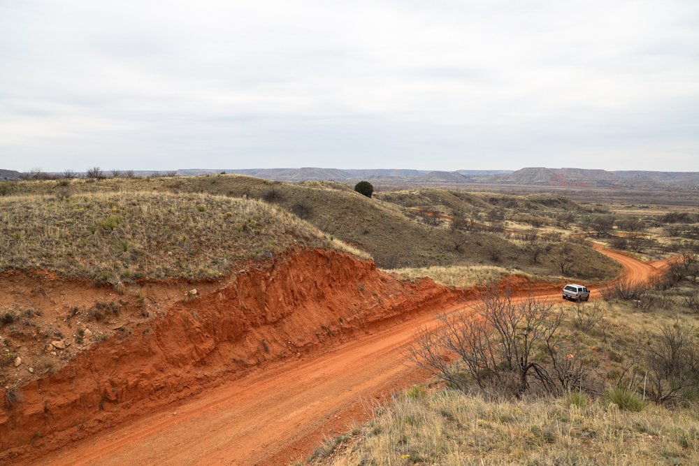 A truck drives down a red dirt road with short grasses on either side