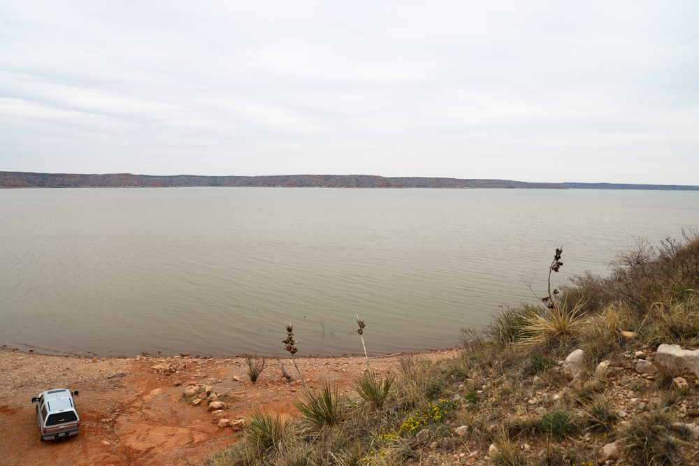 A large blue gray lake in front of red brown soil with a few people and vehicles recreating