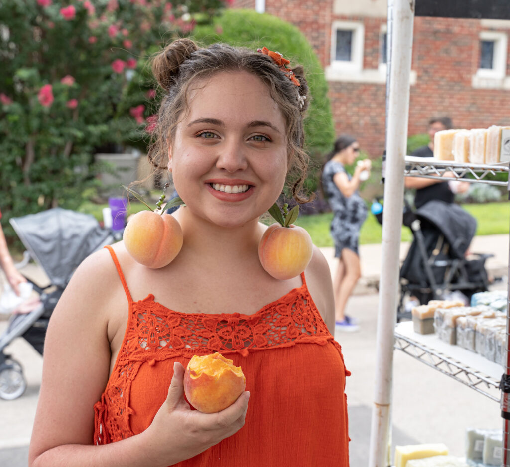 A young women, smiling and holding a peach, wears dangling peach earrings. Photo by Randy Cutshall, courtesy of Weatherford Chamber of Commerce. 