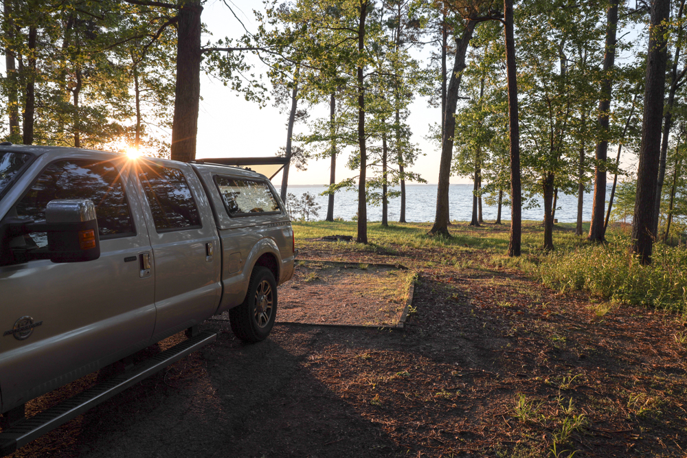 A truck parked with the topper open at the edge of water with tall trees