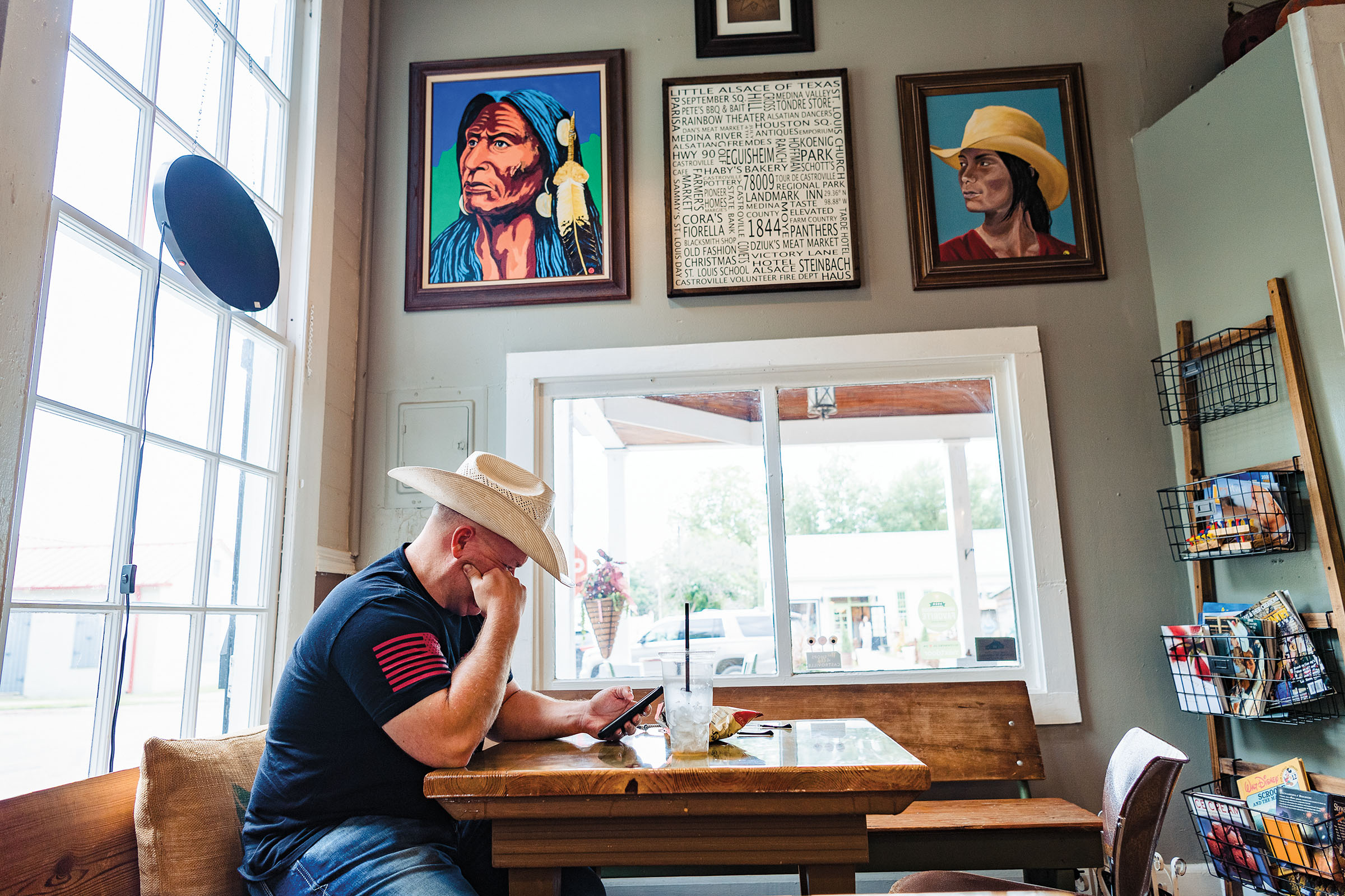 A man sits at a table looking at his phone beneath a wall of portraits