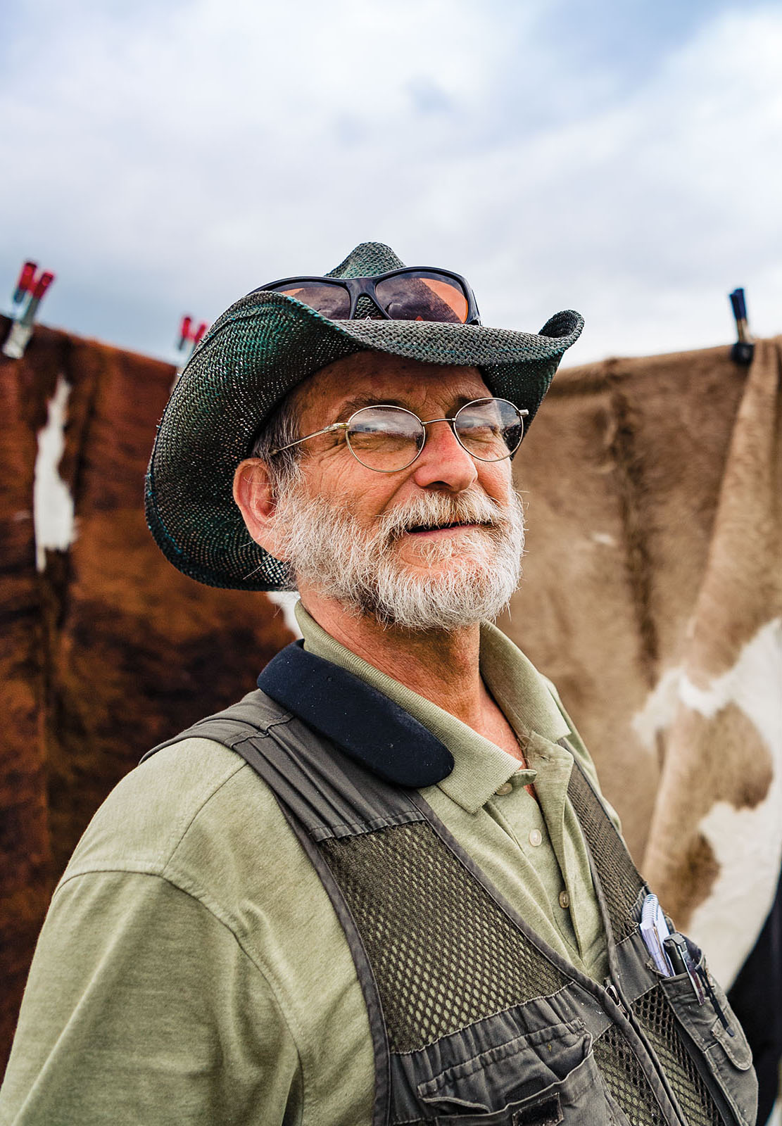 A man in a floppy hat and vest stands in front of a strung-up hide