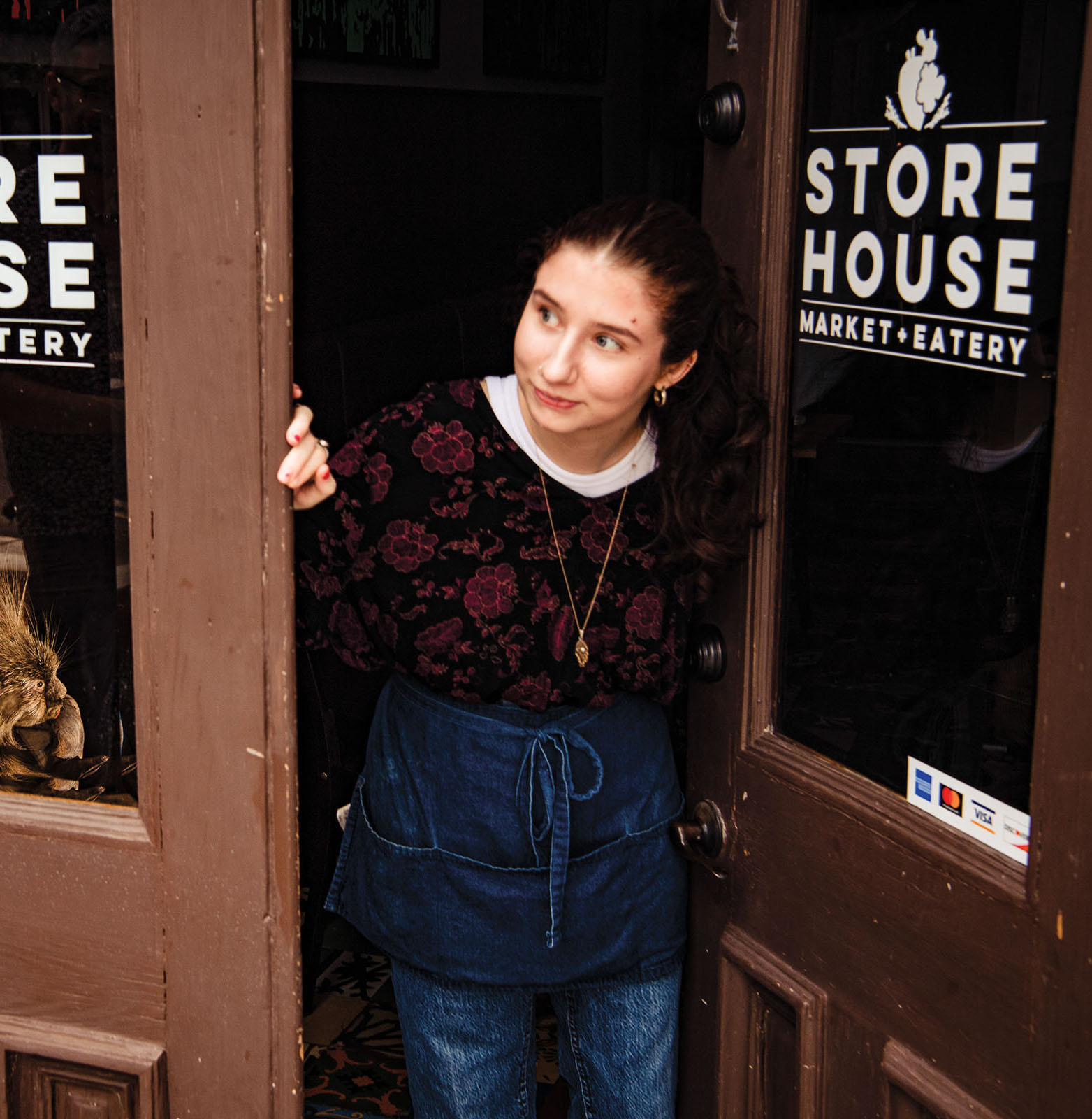 A woman looks out of a brown-painted wood door reading Store House Market + Eatery