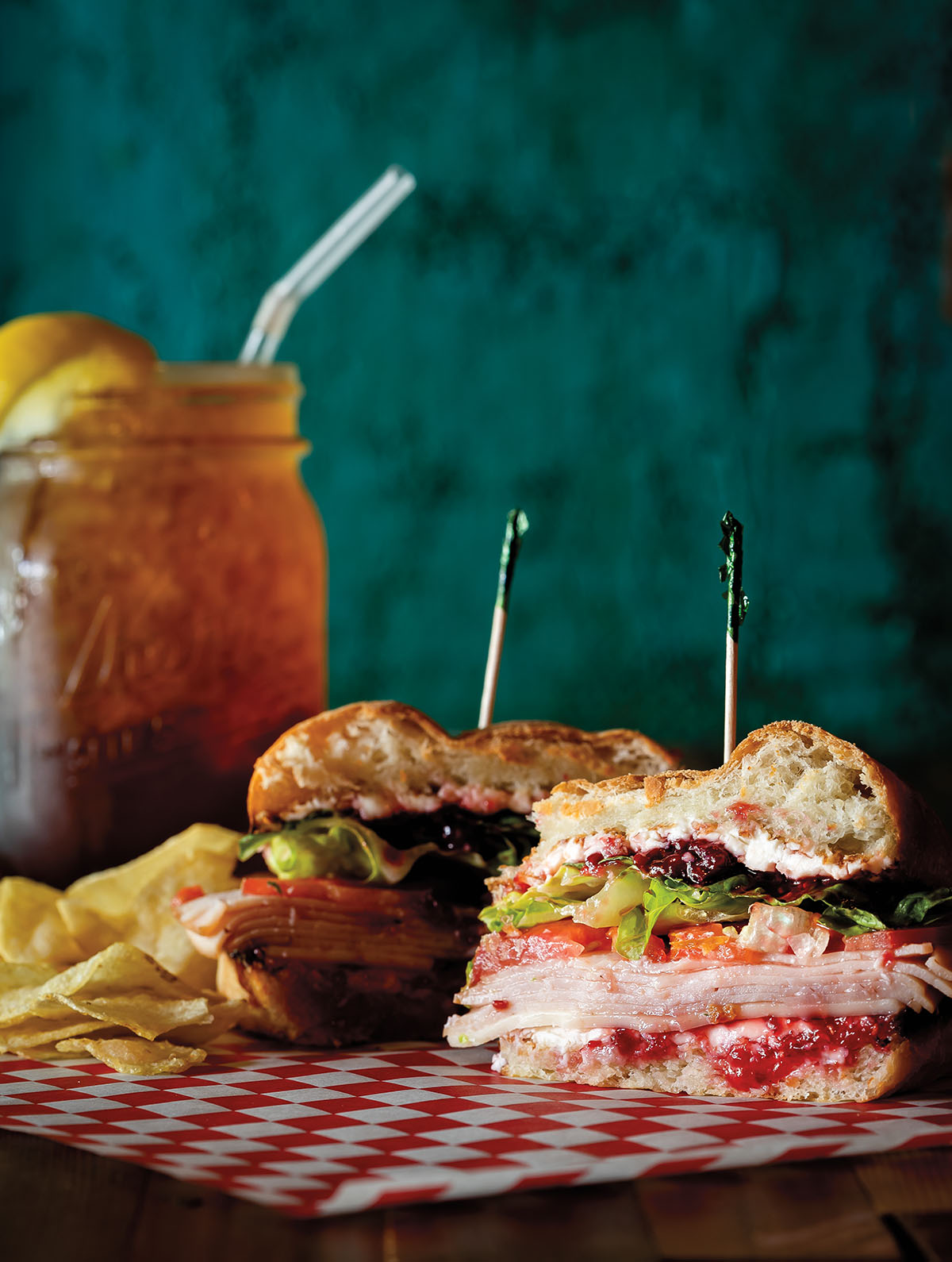 A sandwich stacked high on a red-checkered paper liner in front of chips and iced tea