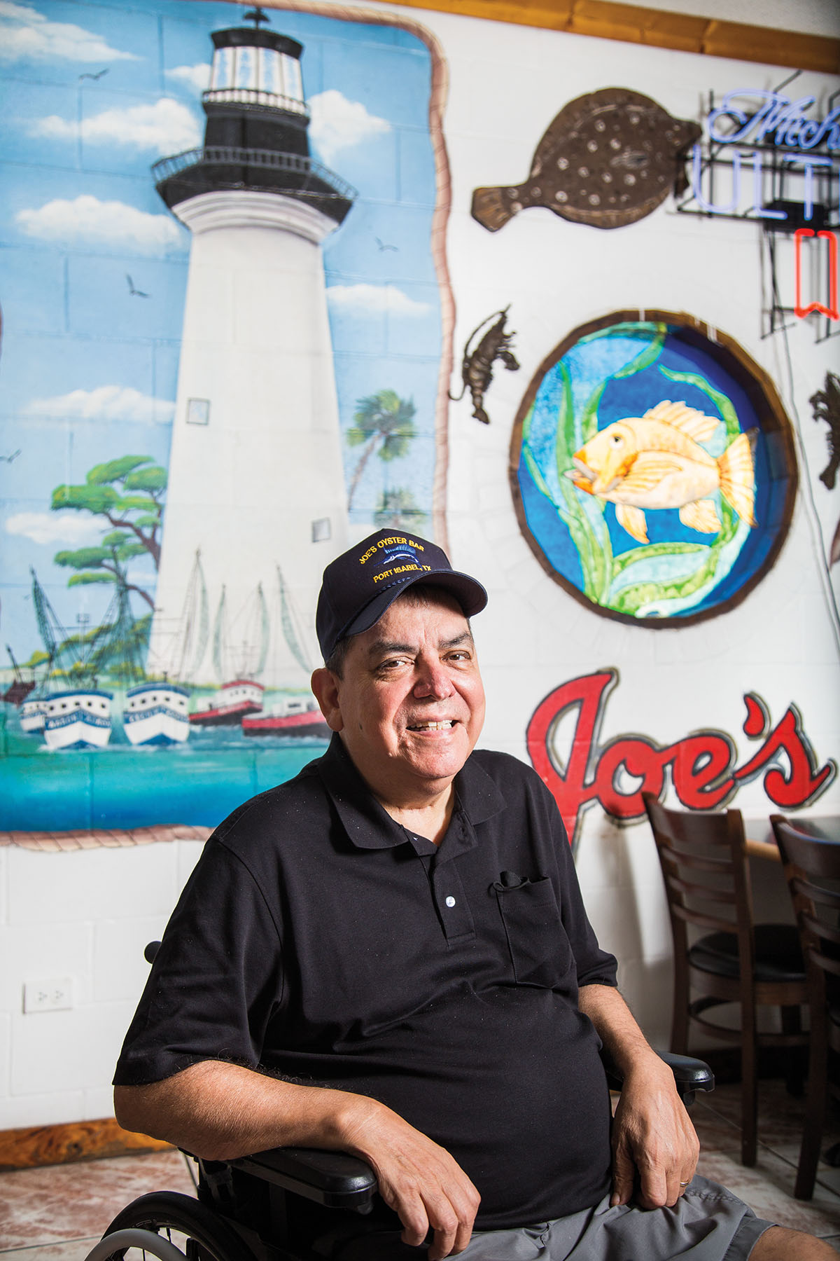 A man in a black shirt and ball cap sits in front of a mural painted lighthouse