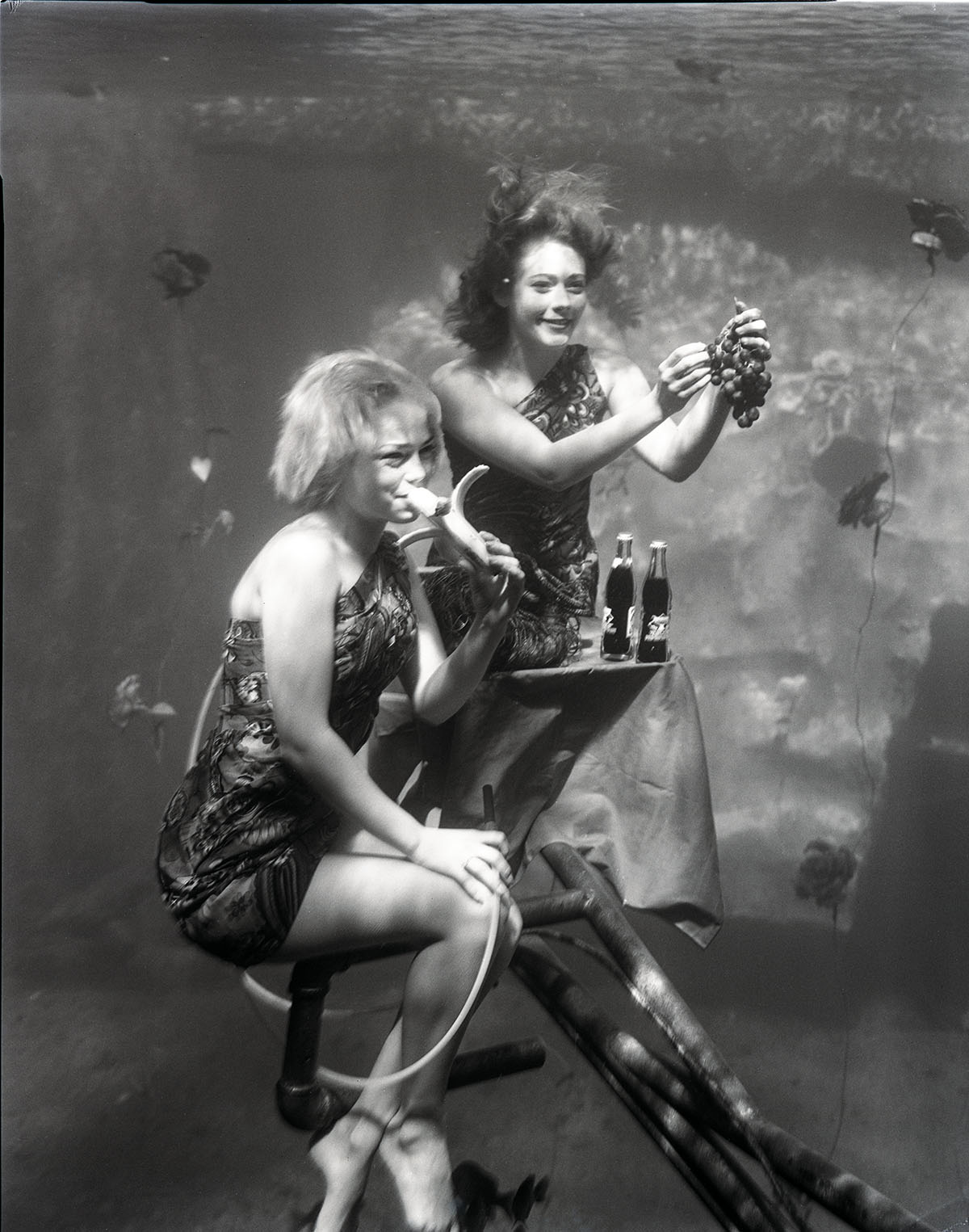 Two women perform underwater surrounded by bubbles in a black and white photograph