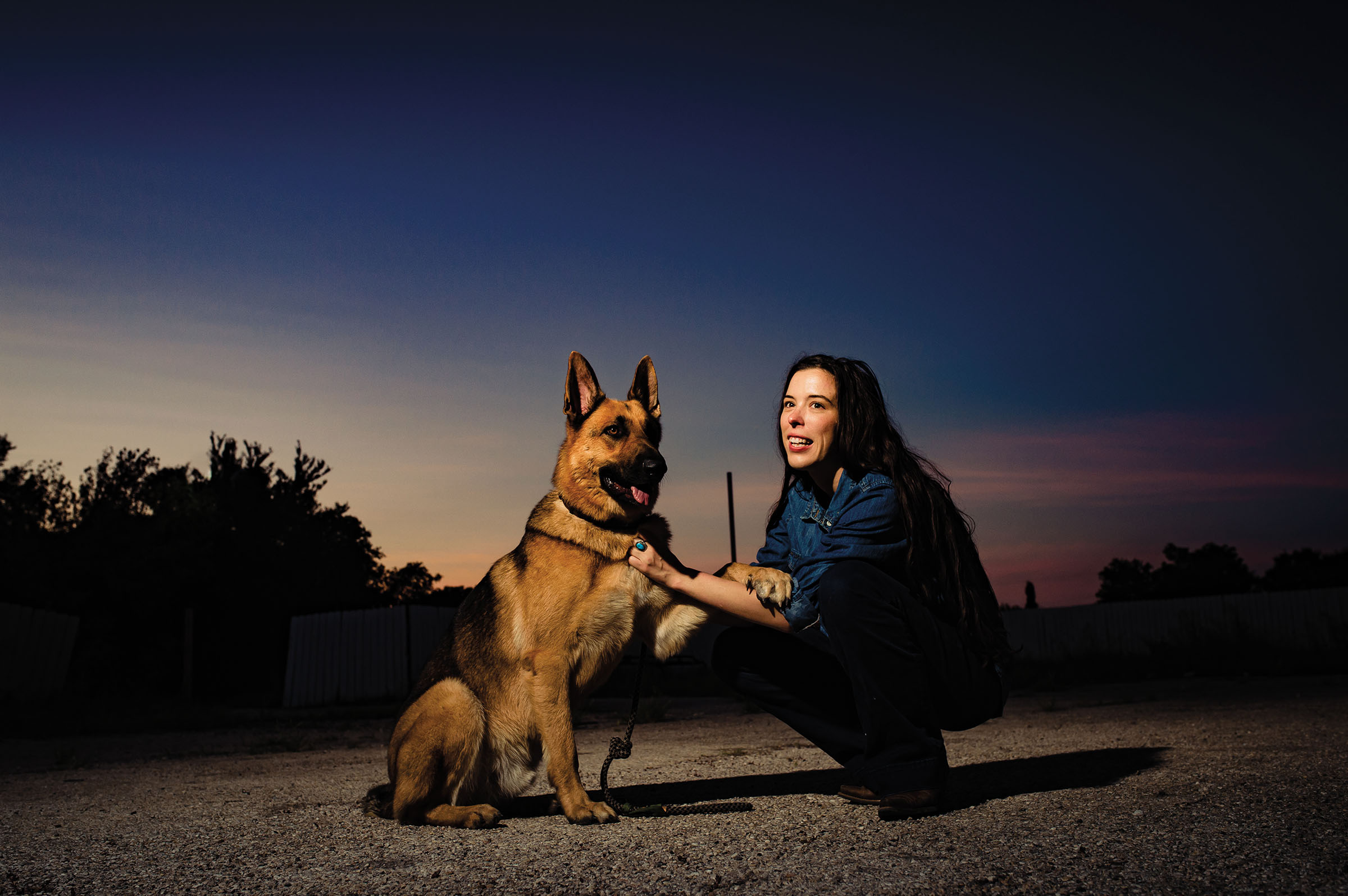 A woman in a blue shirt squats down in the dark next to a german shepherd