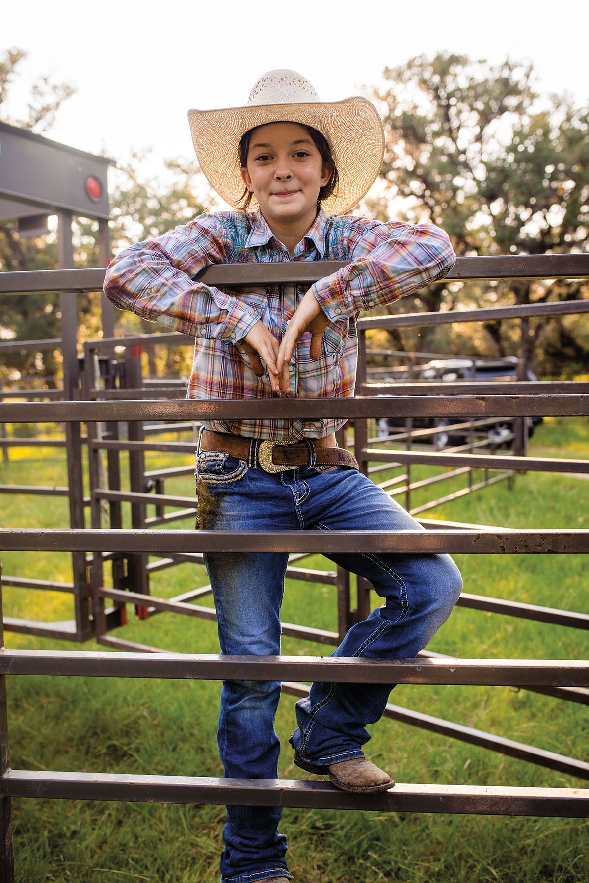 A young woman in a cowboy hat stands with her arms draped over a fence with a slight smile