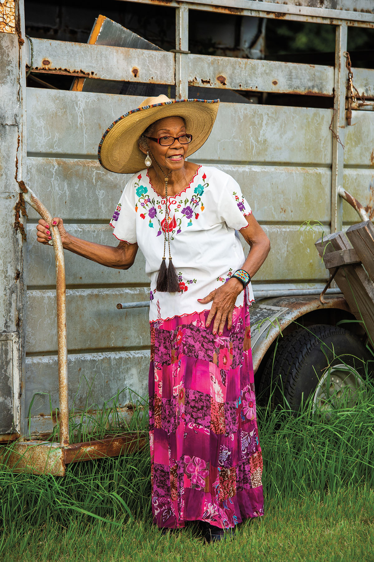 A woman in a hat, white top and brightly colored skirt leans against a metal horse trailer