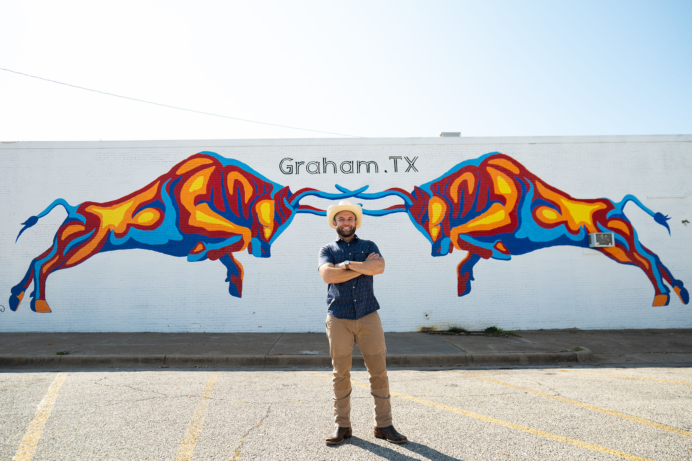 A man in a blue shirt and small cowboy hat stands in front of a mural of two bulls reading Graham, TX