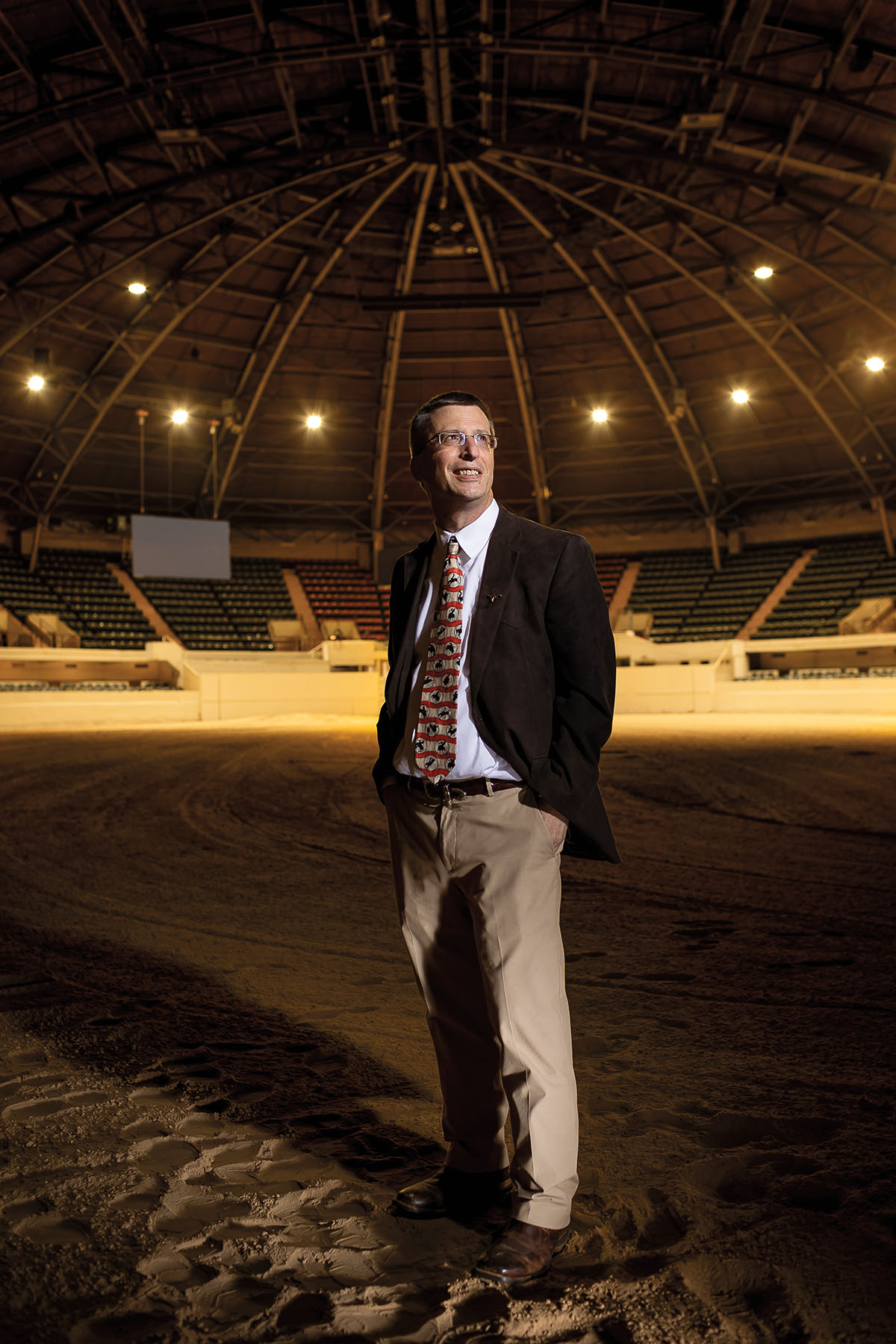 A man in a black blazer, tie and khaki pants stands in a large arena