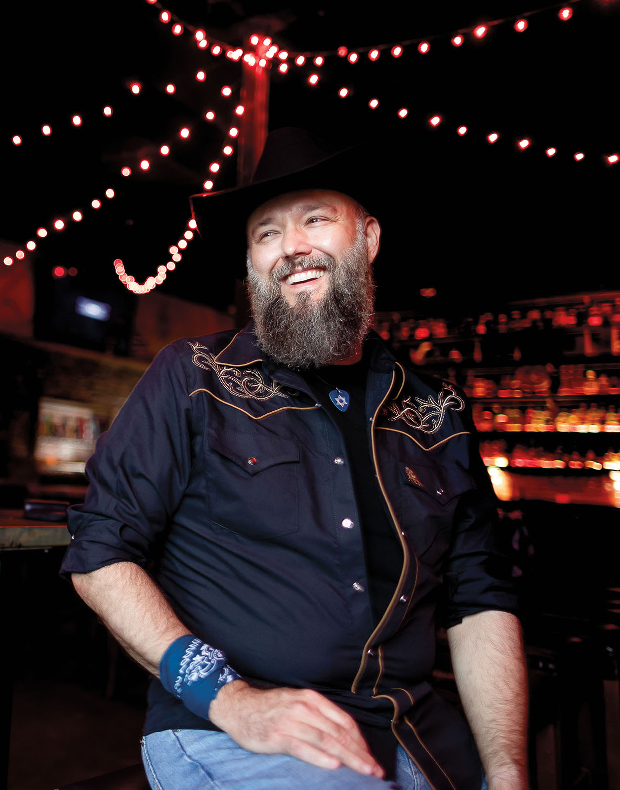 A man in a black cowboy hat and sequined shirt smiles under string lights