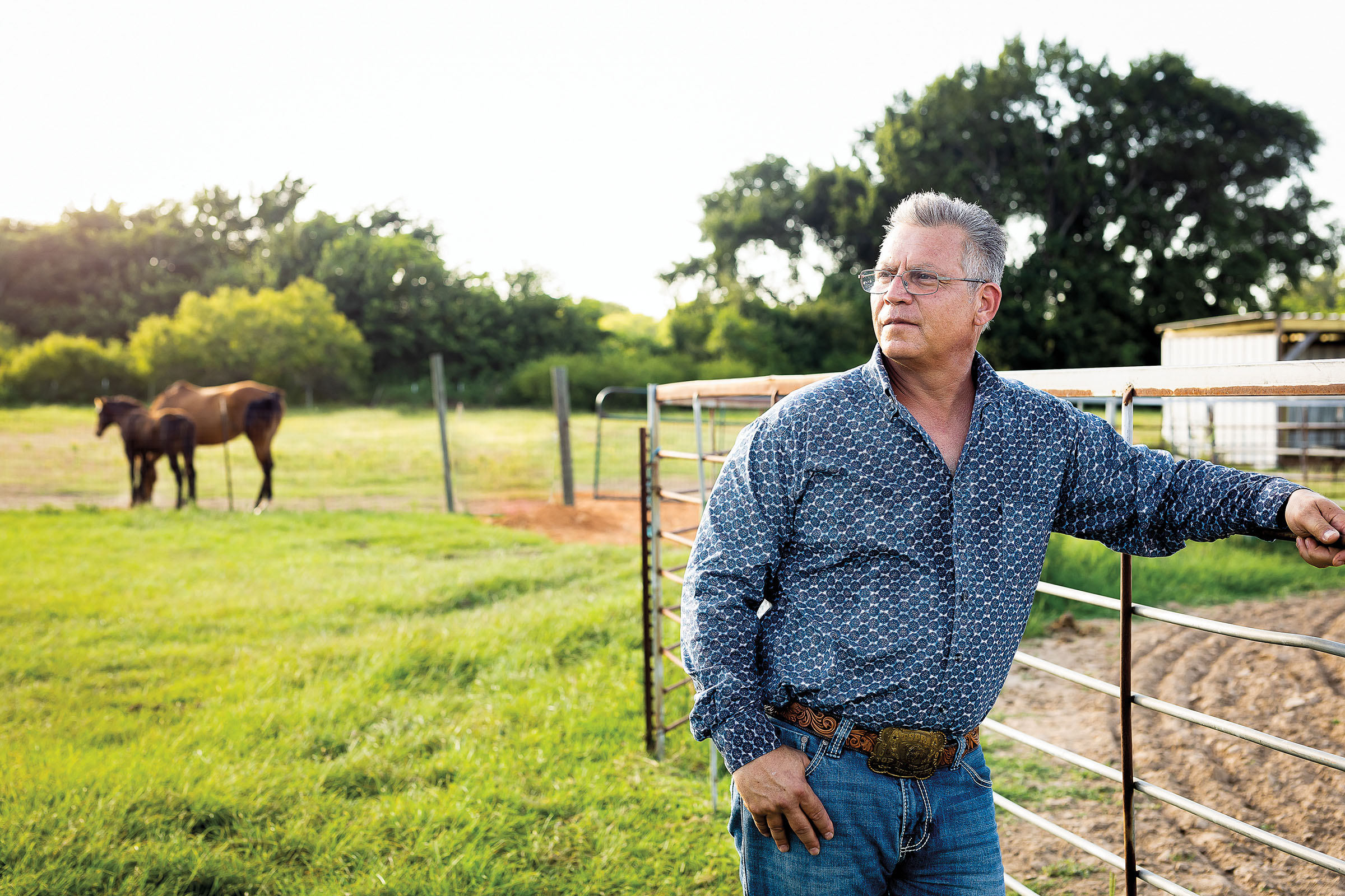 A man in a blue shirt and blue jeans looks off into the distance with his hand in his pocket - standing in a large green pasture