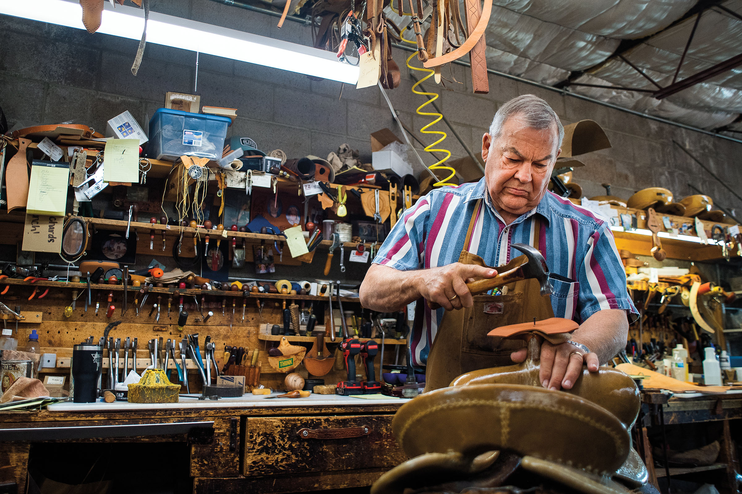 A man in a striped shirt works on a saddle in a busy shop