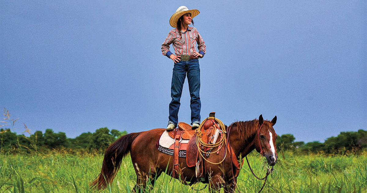 A woman in a brightly-colored shirt and cowboy hat stands on top of a brown horse in a green field