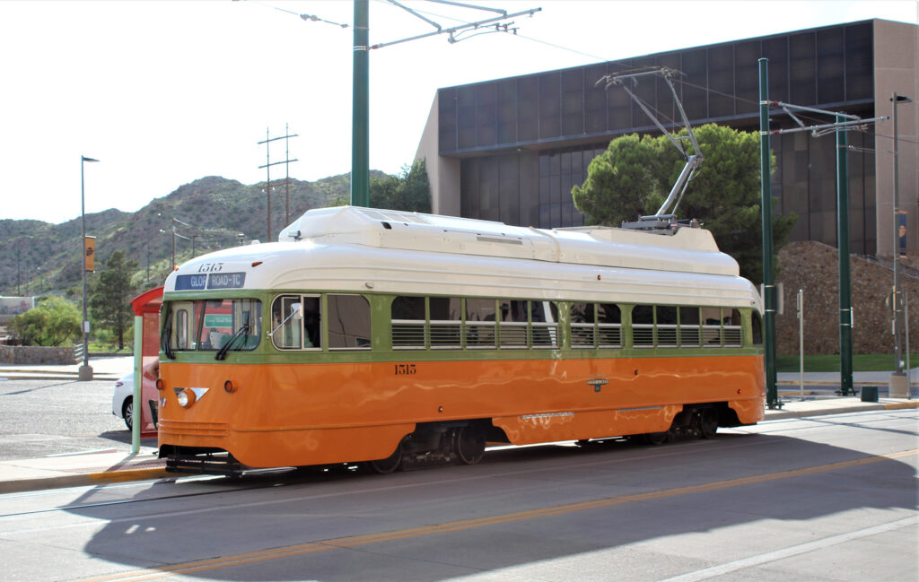 An orange and white electric streetcar stops along the tracks with a modern-day building and El Paso's mountains in the background. Roberto José Andrade Franco. 