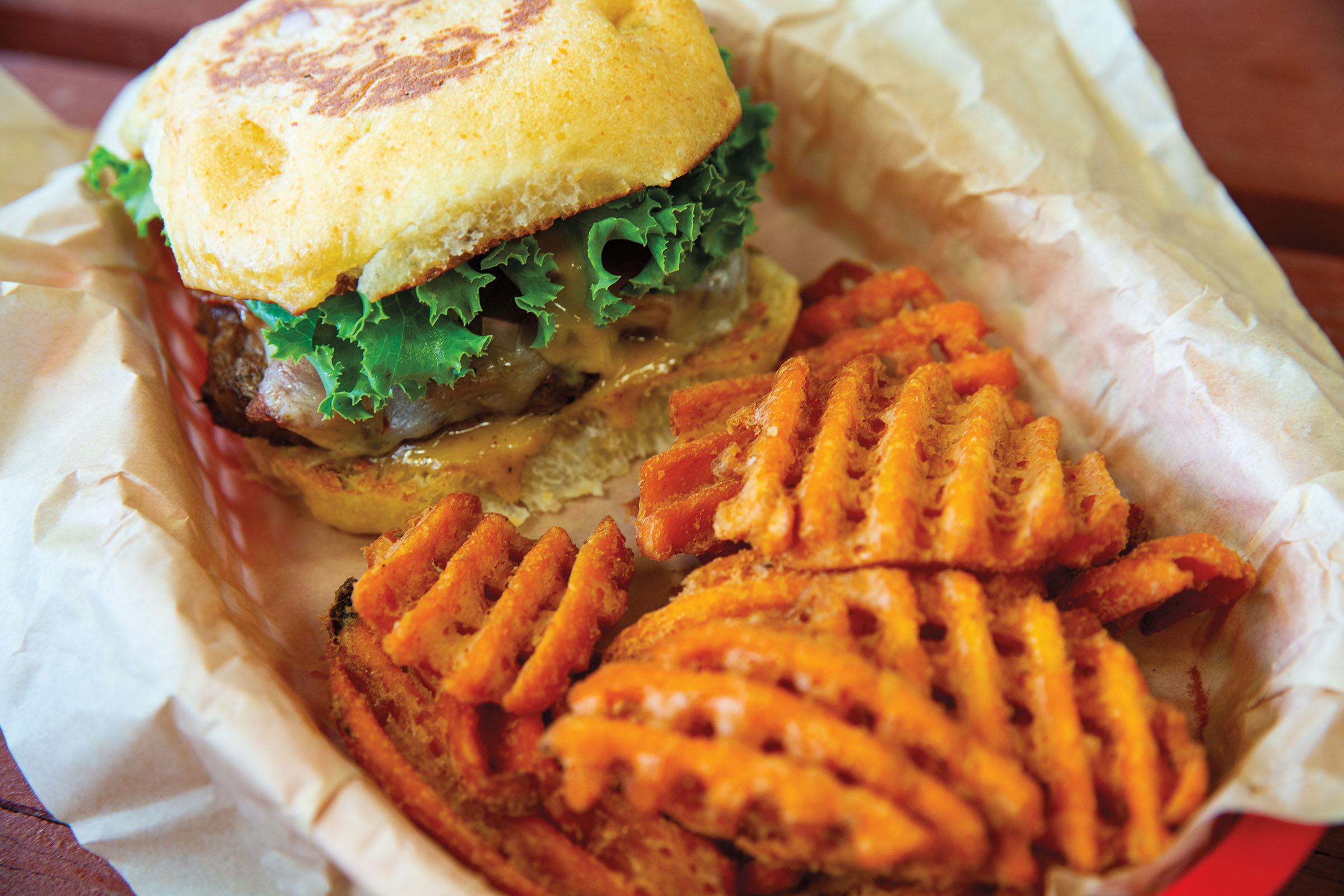 A basket of food containing waffle-cut sweet potato fries, a burger on a yellow bun with a large piece of lettuce and melting cheese