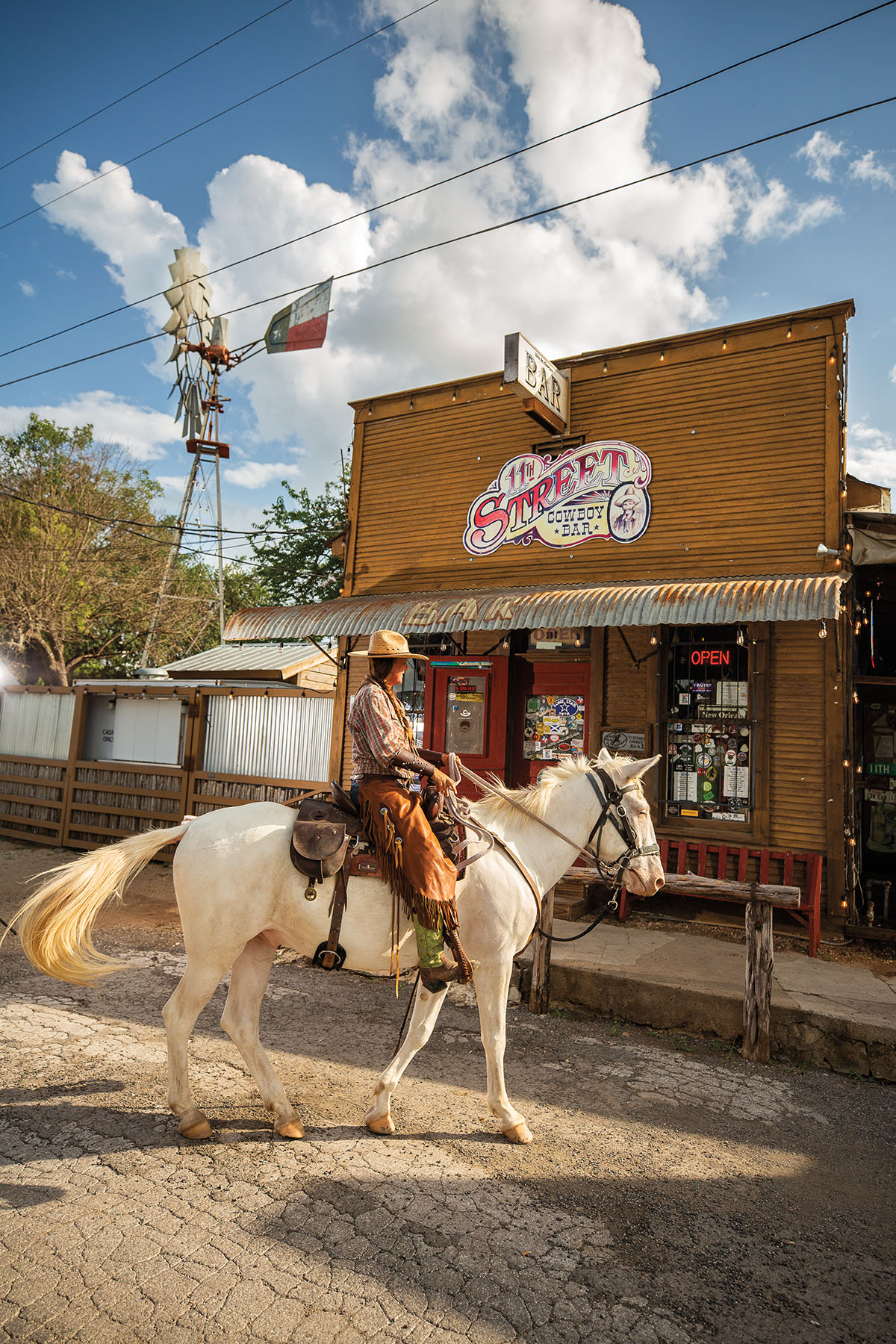 A woman in a tan cowboy hat and brown chaps rides a white horse in front of a wooden saloon reading "street cowboy bar"