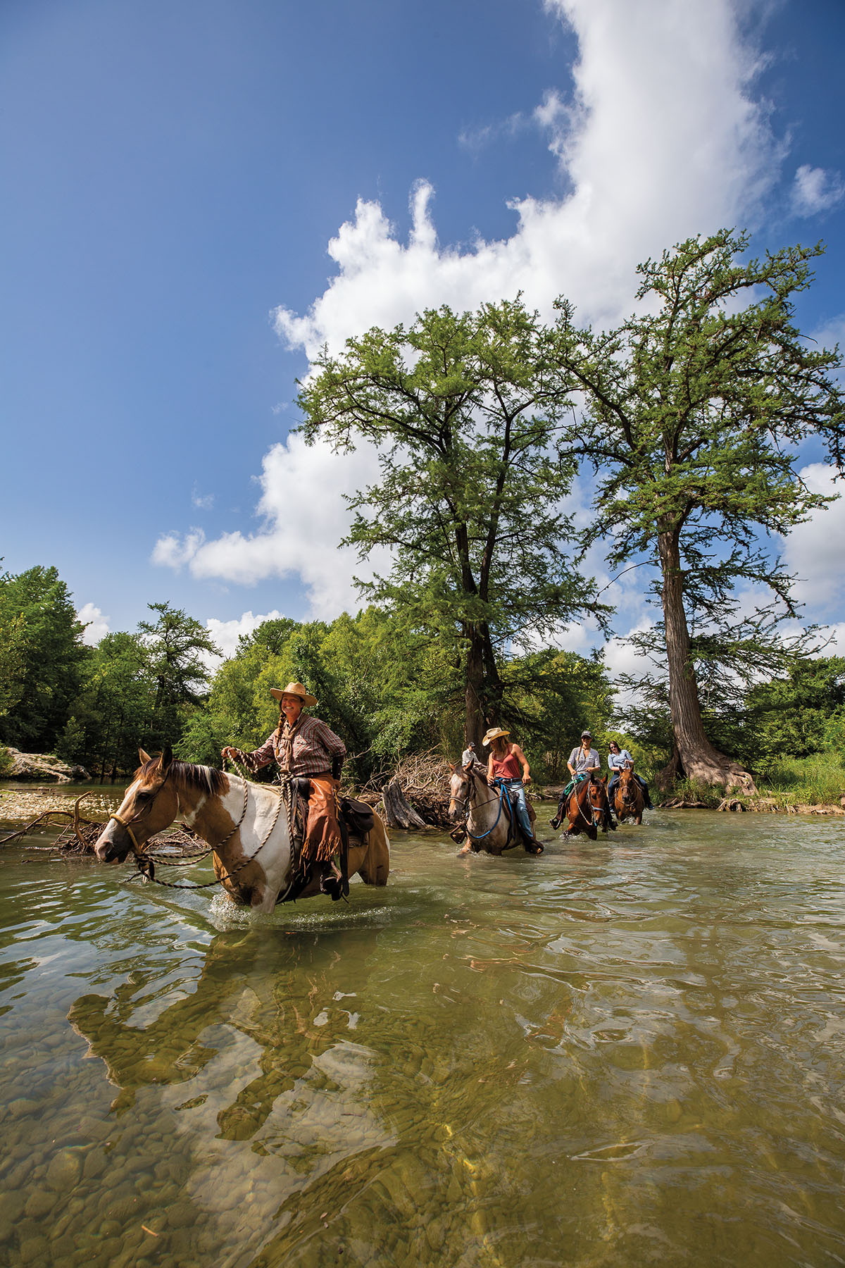 A group of people smile as they ride horses through water under bright blue sky