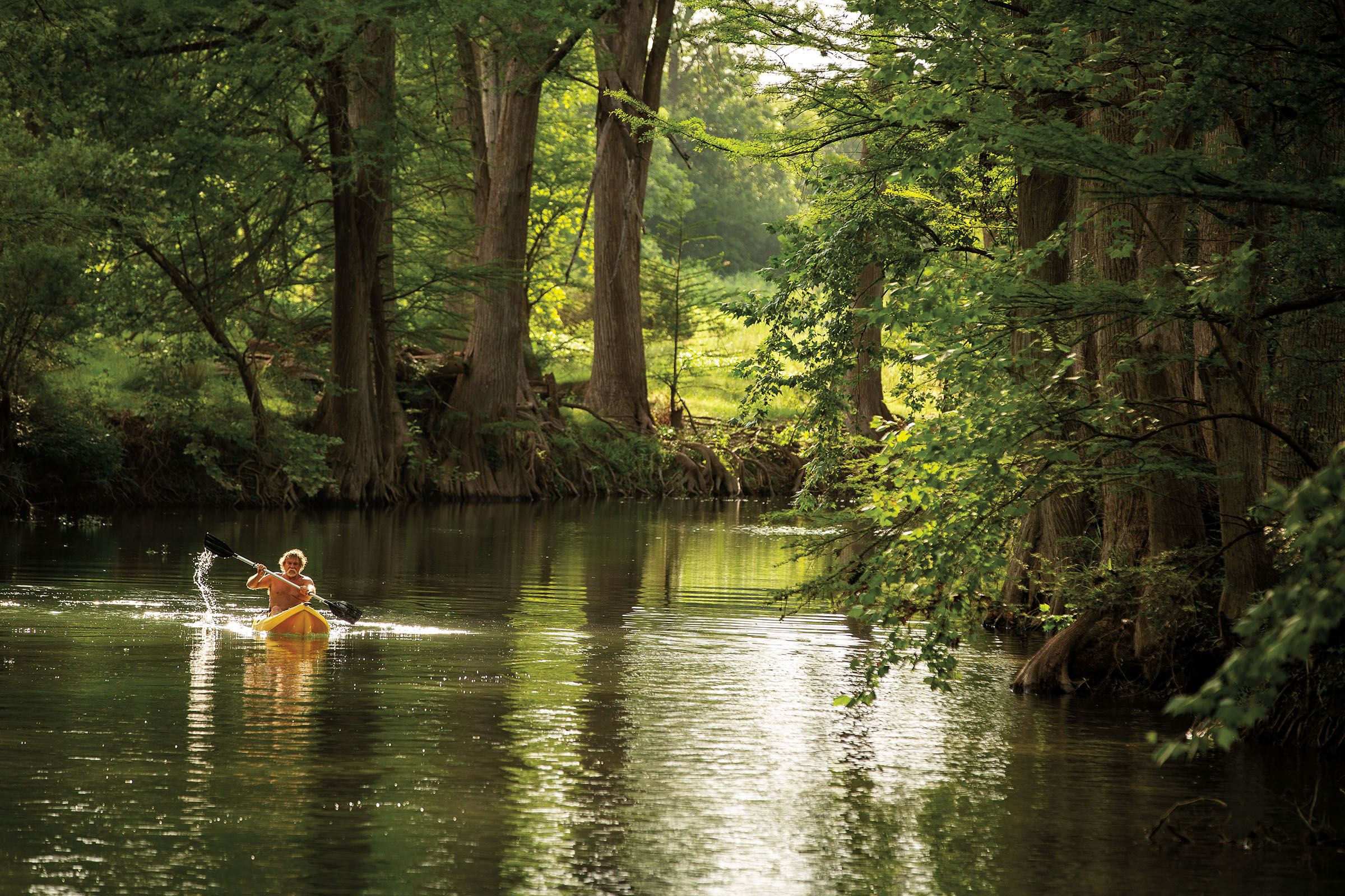 A shirtless man in a yellow kayak paddles down a lush green waterway