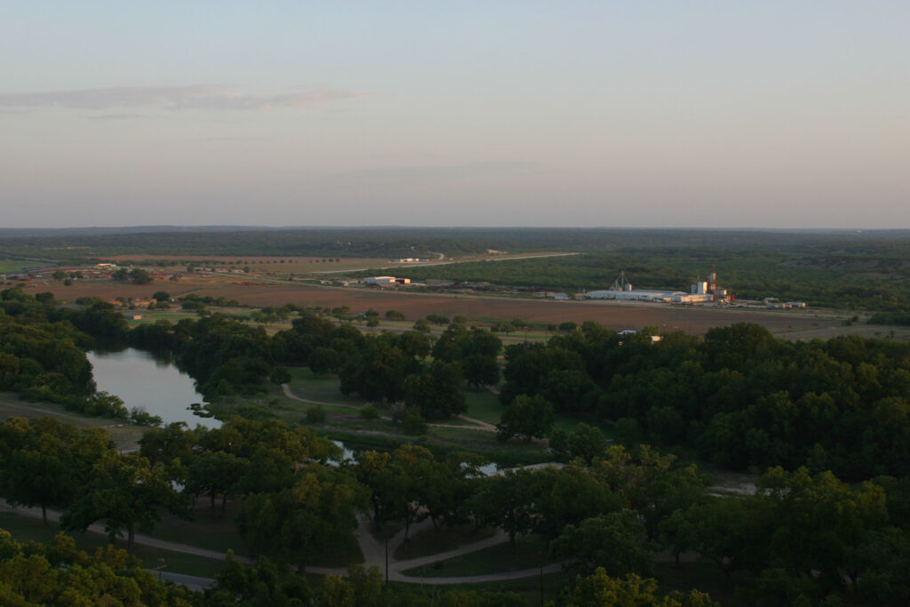 An overhead shot of the small town of Menard, with sky overhead and fields, roads, a river, and trees in the foreground. 